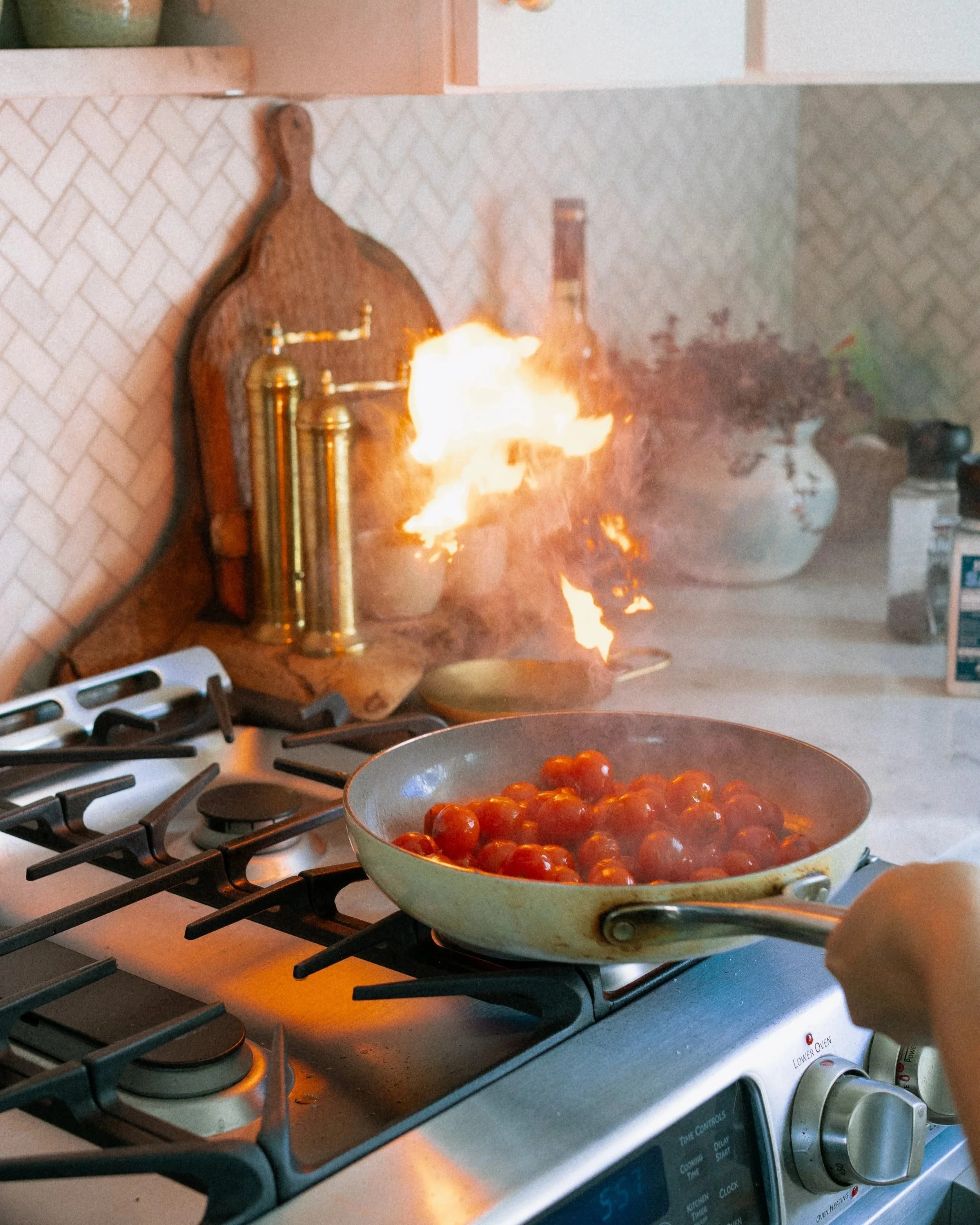 A stove with a frying pan of cherry tomatoes on one burner and a fire explosion on the backsplash near the wall.