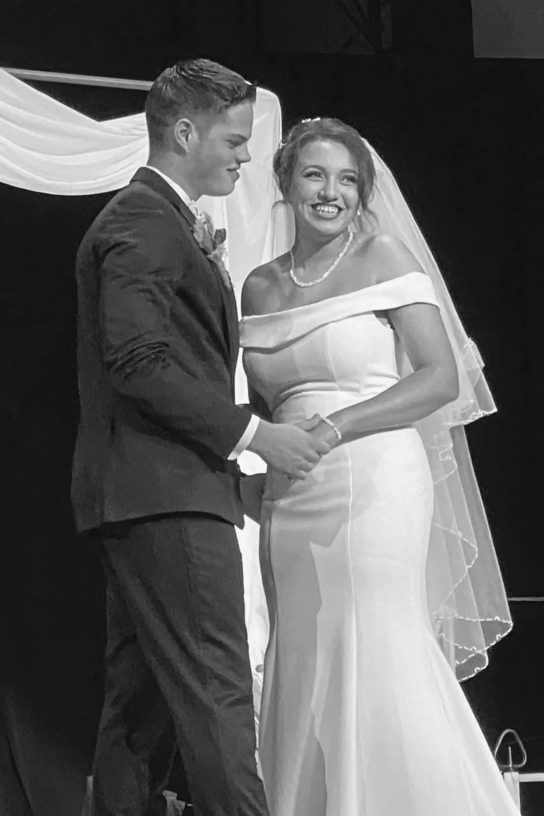 A bride and groom holding hands during their wedding ceremony, smiling at each other. The bride wears an off-the-shoulder wedding gown with a veil, and the groom wears a dark suit with a boutonniere.