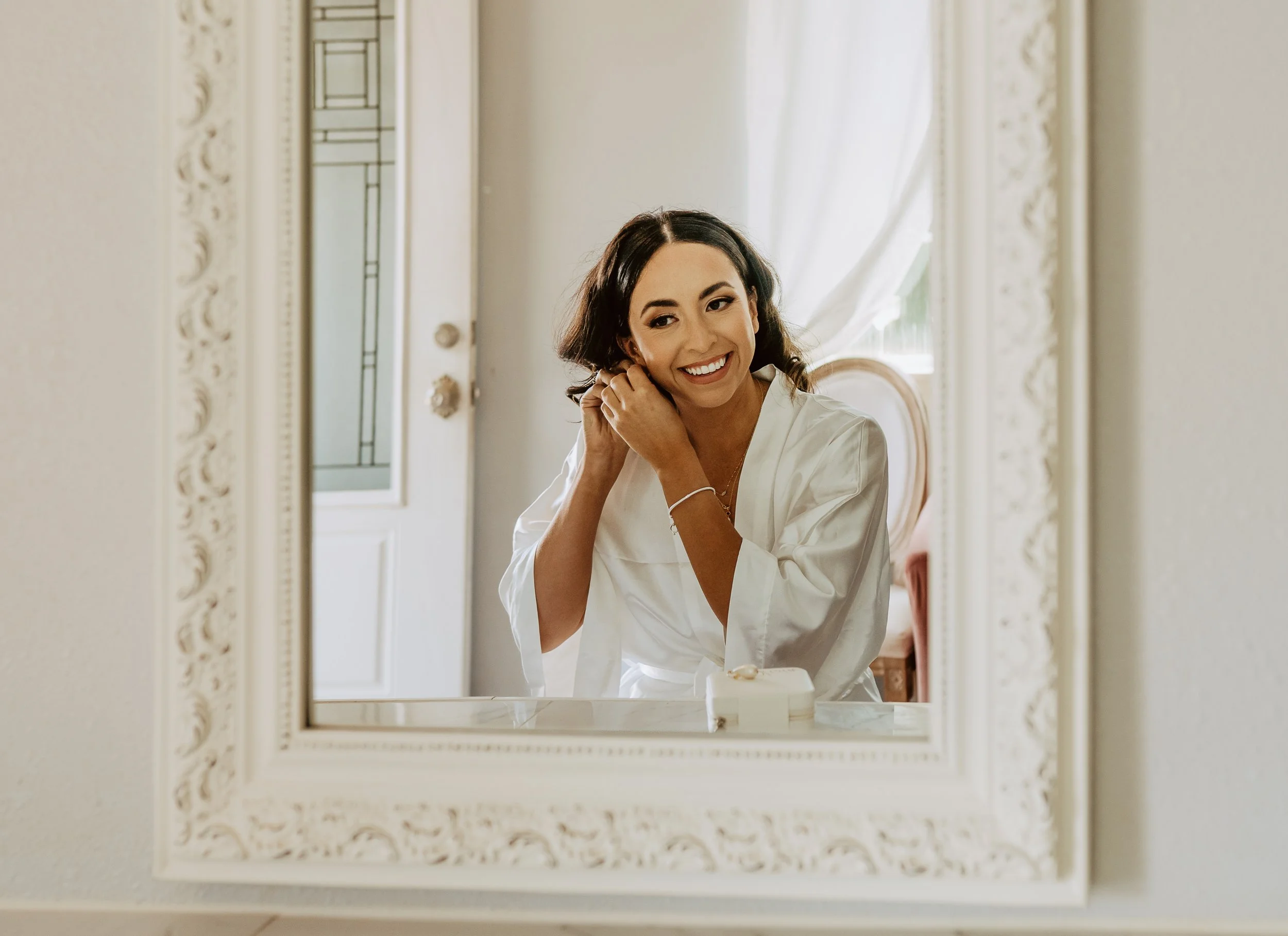 A woman with dark hair and a white satin robe smiling and adjusting her earring, seen through a decorative mirror with a white ornate frame, in a bright room with white curtains and a window.