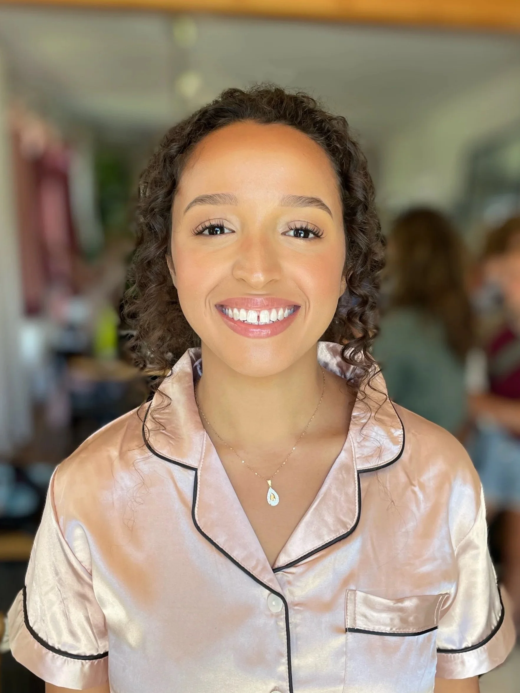 A young woman with curly hair smiling, wearing light pink satin pajamas with black piping, and a delicate gold necklace with a drop-shaped pendant, in a cozy, blurred indoor setting.