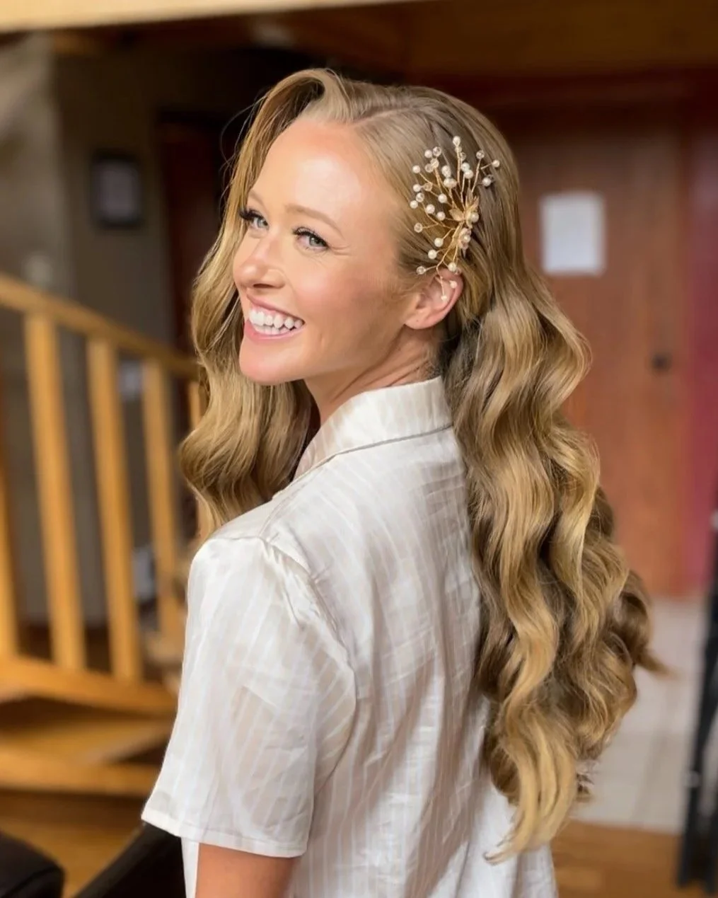 A woman with long, wavy blonde hair wearing a decorative hairpiece with pearls and gold branches, smiling and looking over her shoulder indoors.