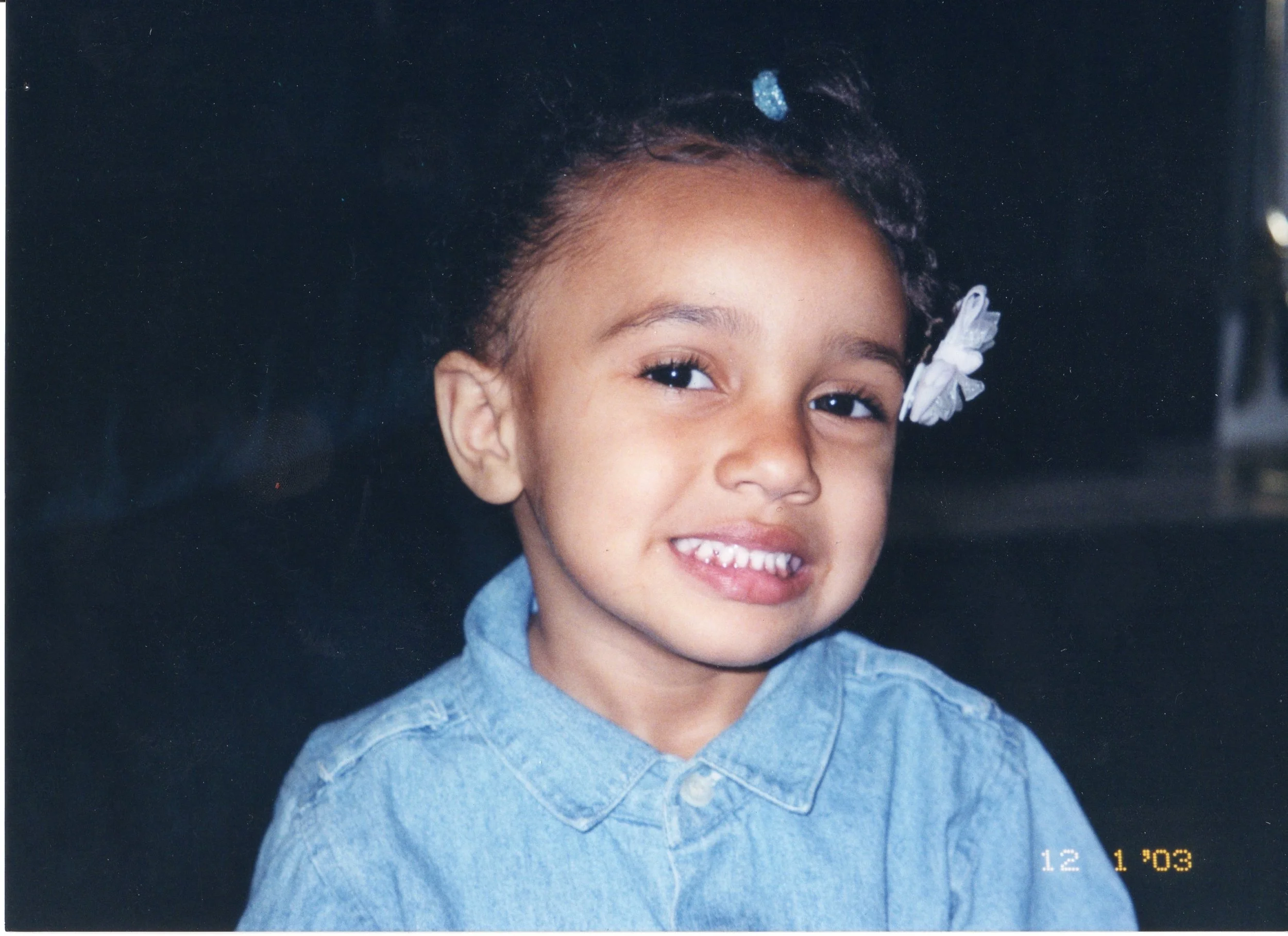 A young girl with curly hair tied with a white bow, wearing a blue denim shirt, smiling at the camera.