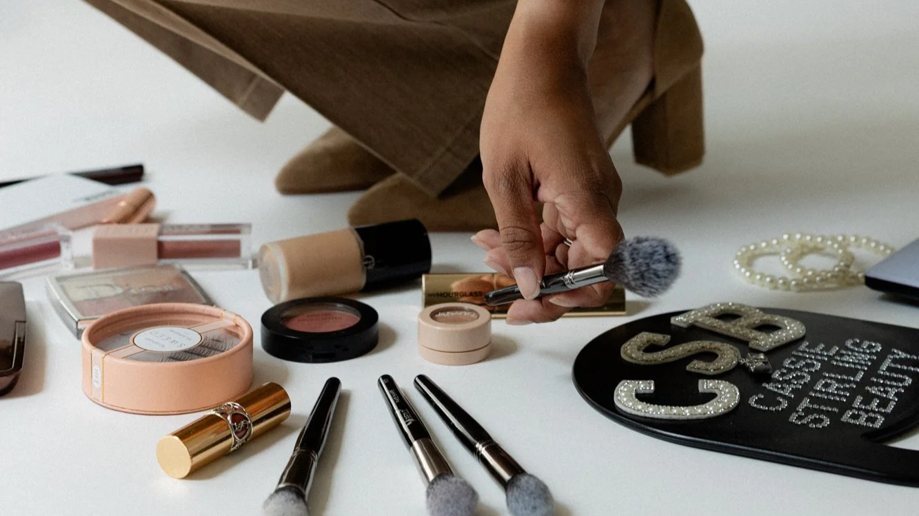 Person's hand holding a makeup brush above a collection of makeup products, including powders, lip gloss, and brushes, on a white surface.