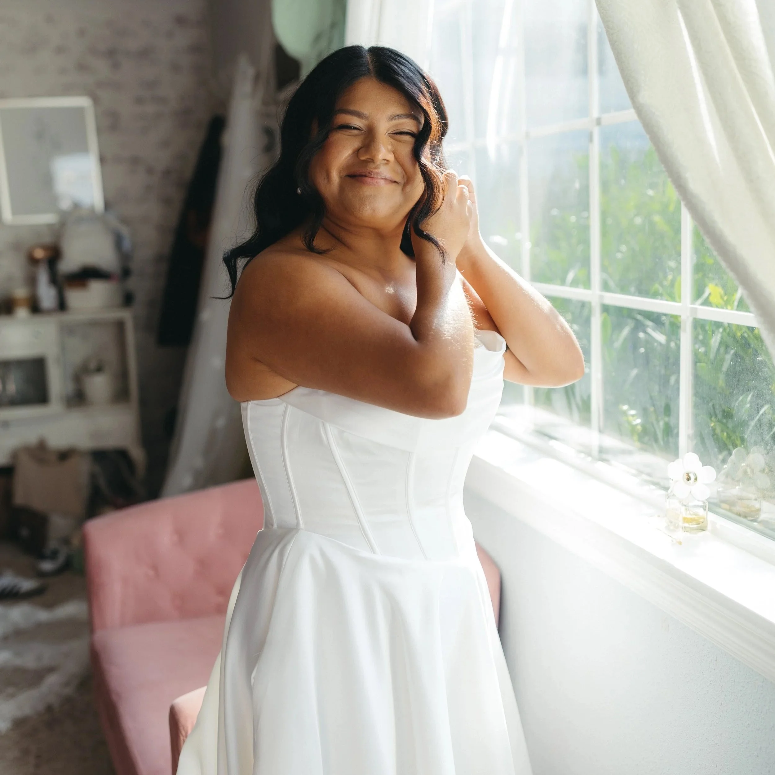 A woman smiling and looking out a window with sunlight streaming in, wearing a white strapless dress, in a cozy room with a pink sofa and shelves in the background.