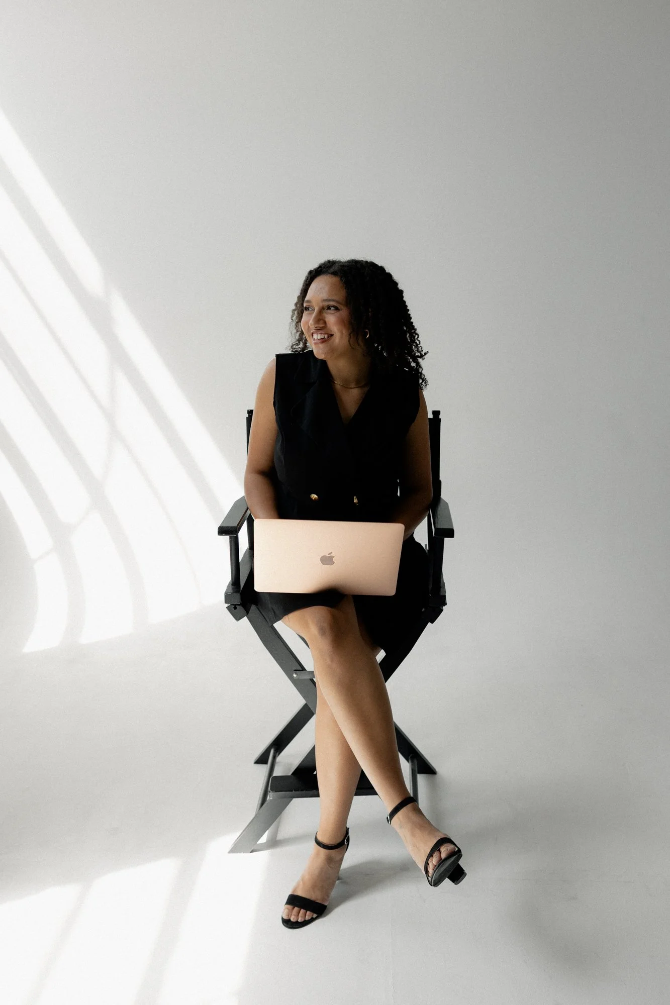 A woman with curly hair sitting on a director's chair, smiling, with a MacBook on her lap, against a plain white background.