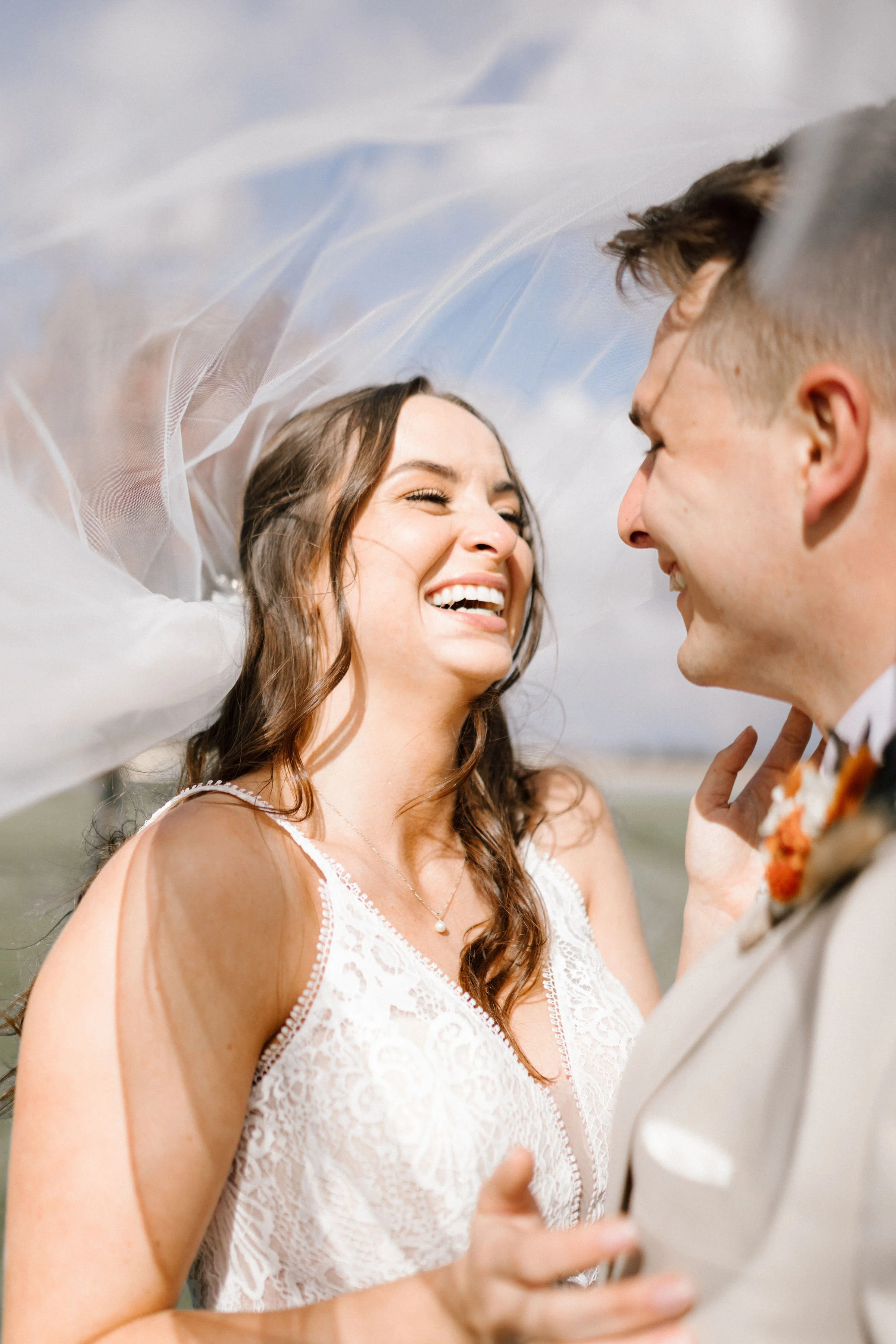 A smiling woman with long brown hair and a pearl necklace looks at a man with short brown hair. They are close, outdoors, with a blue sky and clouds in the background. She is wearing a white lace dress.
