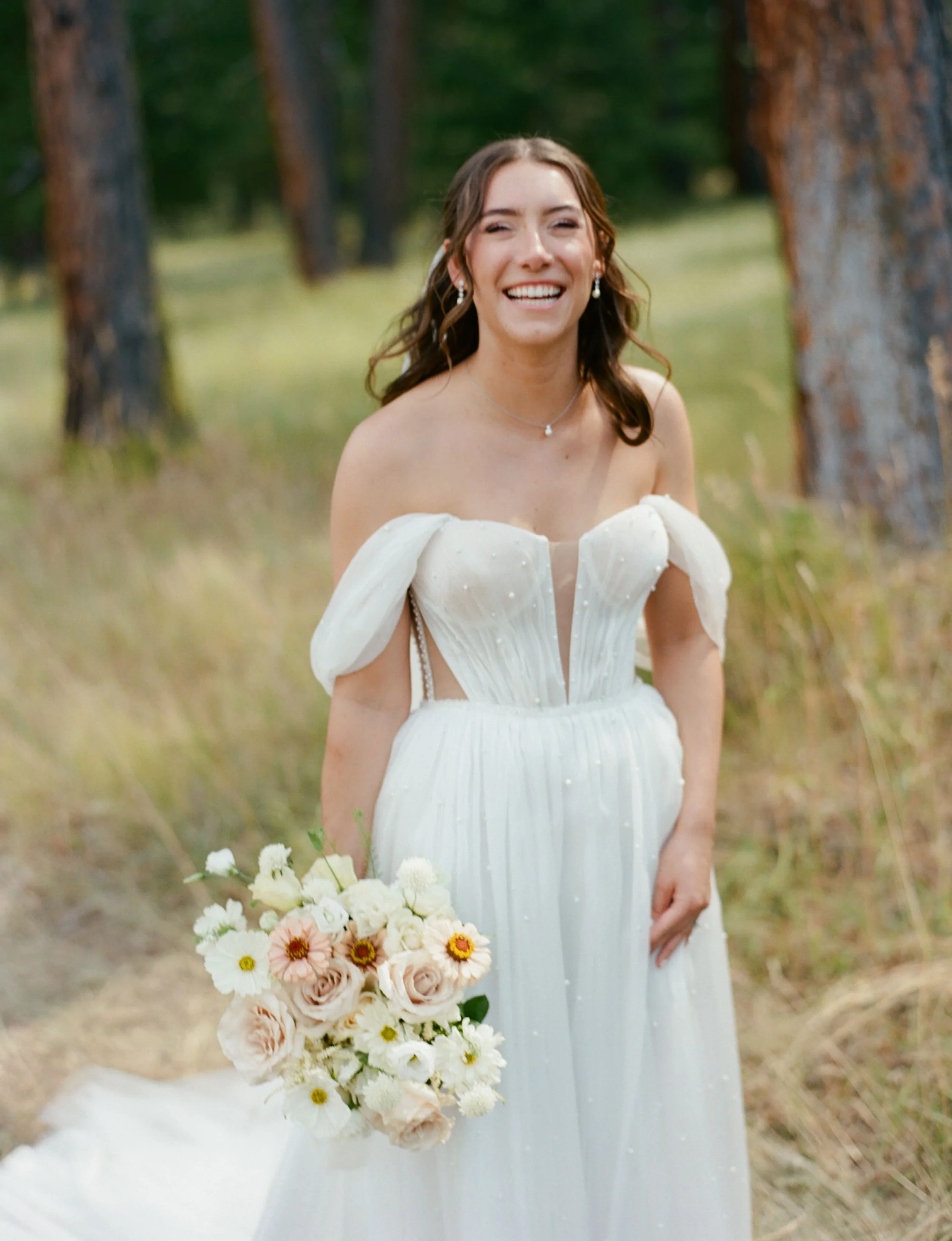A woman in a white off-the-shoulder dress holding a bouquet of white, pink, and peach flowers in an outdoor setting with trees and grass.
