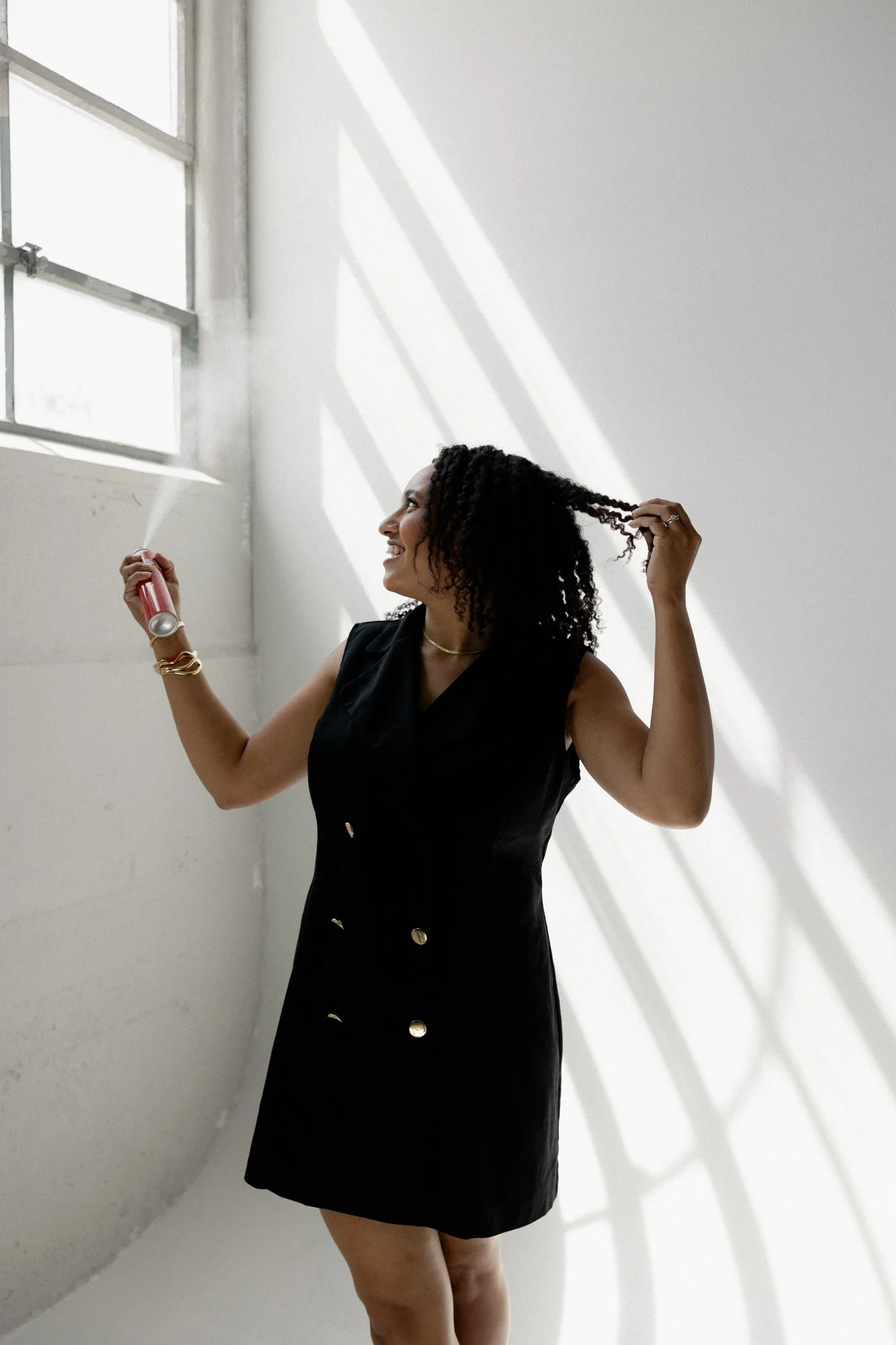 A woman with curly hair wearing a black sleeveless dress, holding a can of soda in one hand and playing with her hair with the other, standing in front of a white wall with sunlight casting shadows from a window.