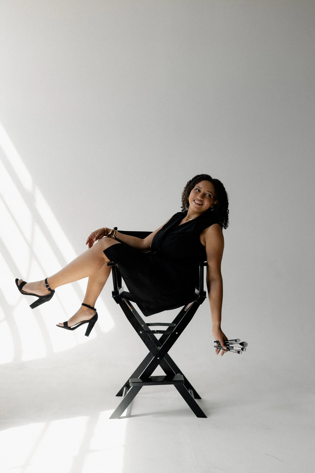 A woman with curly dark hair sitting on a director's chair in front of a plain white background. She is smiling, wearing a black sleeveless dress, platform heels, and jewelry, holding makeup brushes in her right hand, with her left arm resting on her knee.