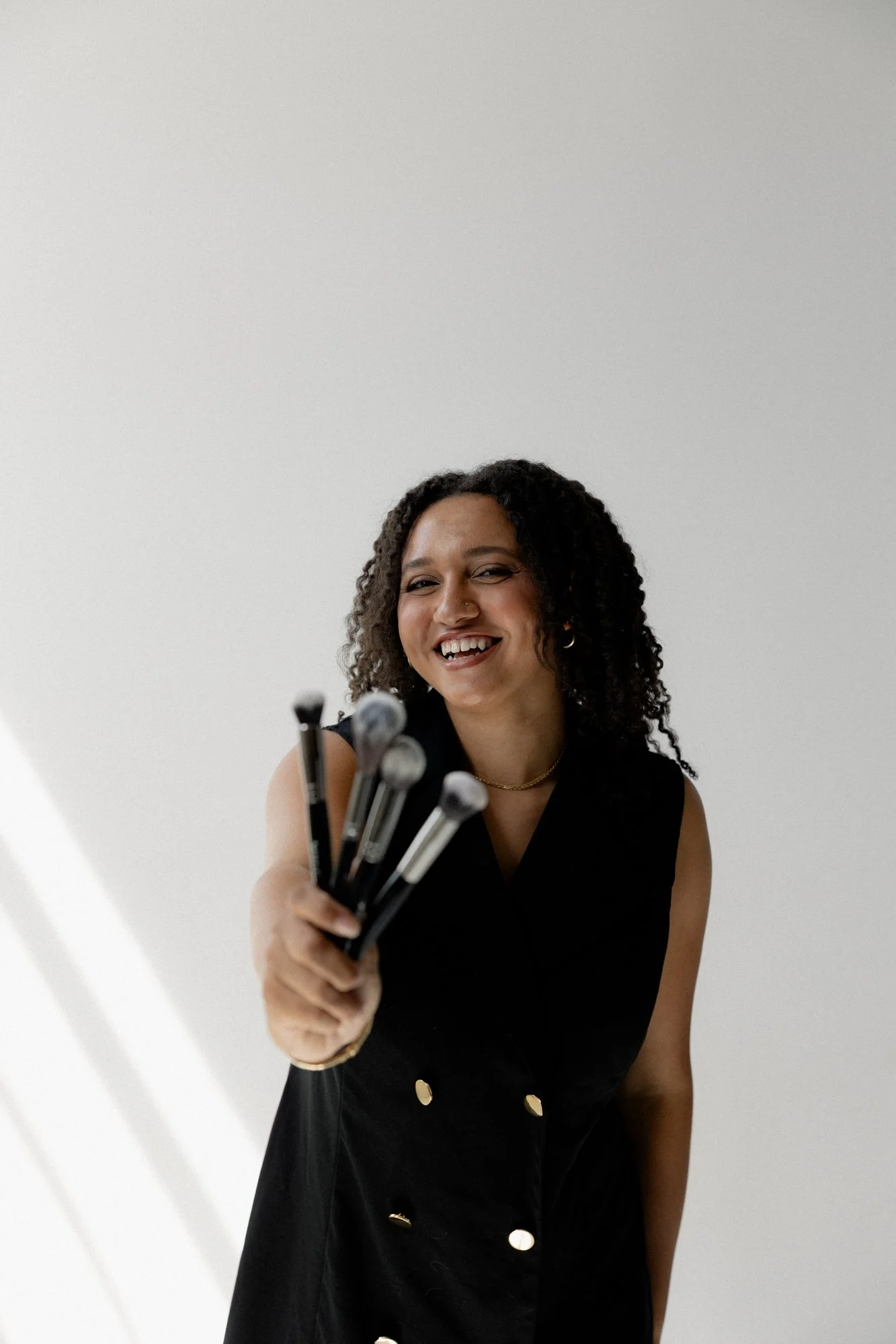 A woman with curly hair wearing a black dress with gold buttons, smiling and holding a set of makeup brushes in front of a plain white wall.