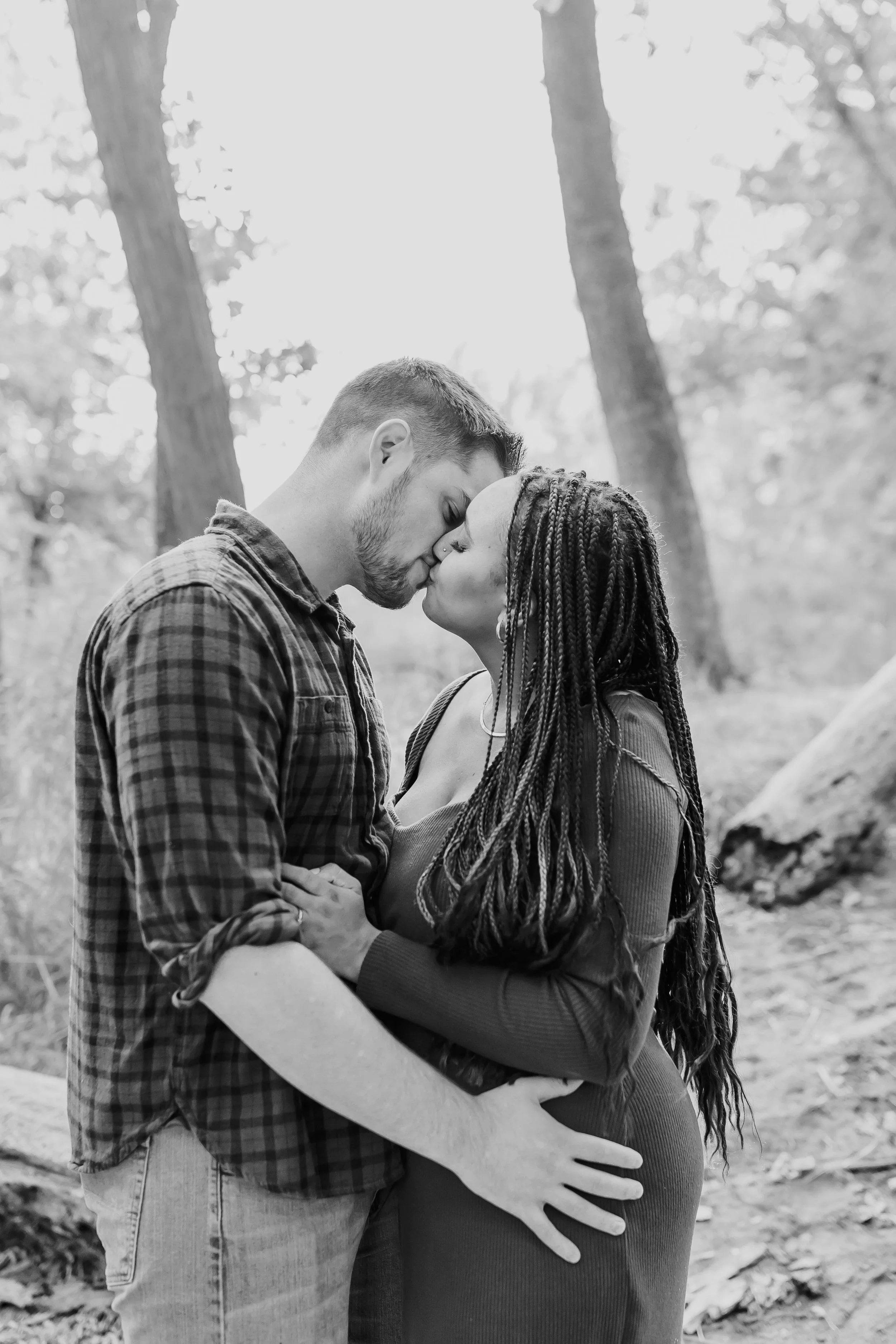 A couple is standing outdoors, nose to nose, sharing a tender moment. The man has short hair and a beard, wearing a plaid shirt, while the woman has long braided hair, wearing a dress. They are surrounded by trees in a natural setting.