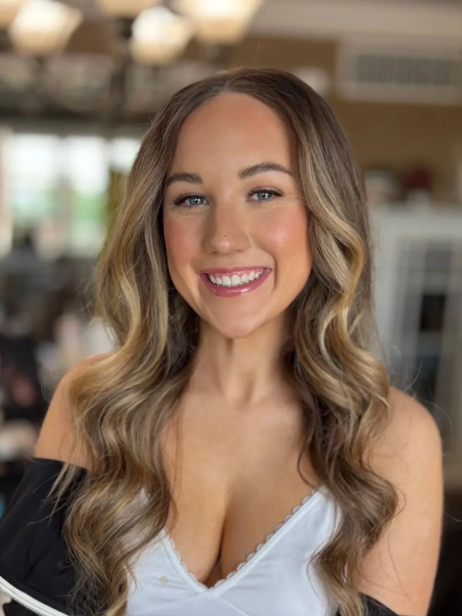 A woman with long wavy blonde hair and blue eyes smiling at the camera, wearing a white top and a black jacket, in a room with warm lighting and blurred background.