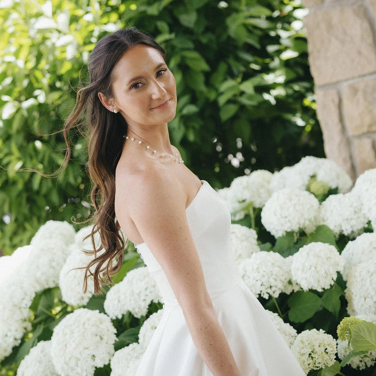 A bride in a white wedding dress standing beside white hydrangea flowers with green leaves and a stone wall in the background.