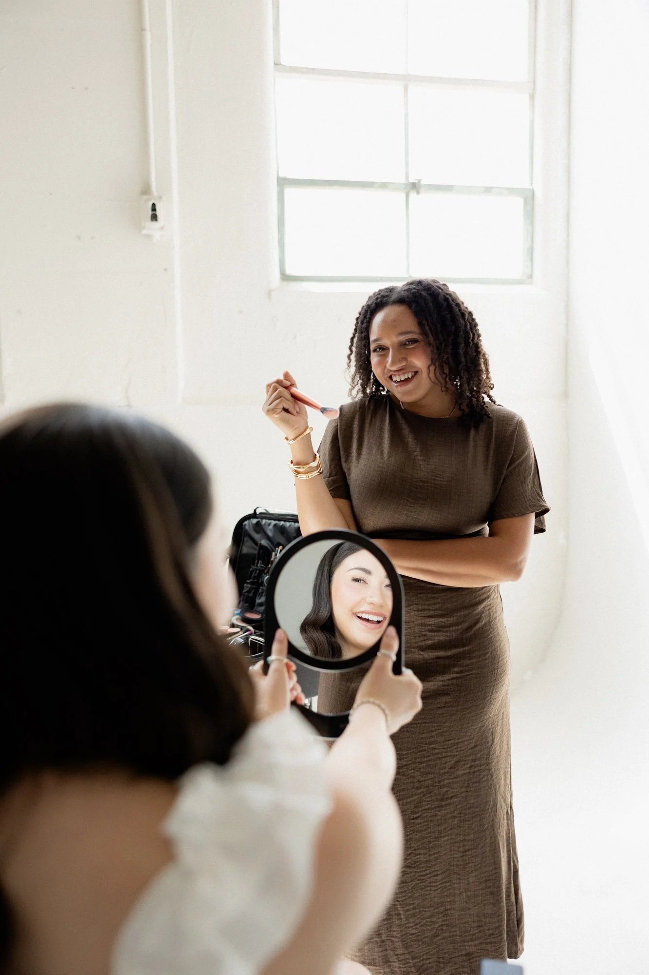 A woman in a brown dress is smiling and holding a makeup brush, while another person is holding a mirror showing her reflection.