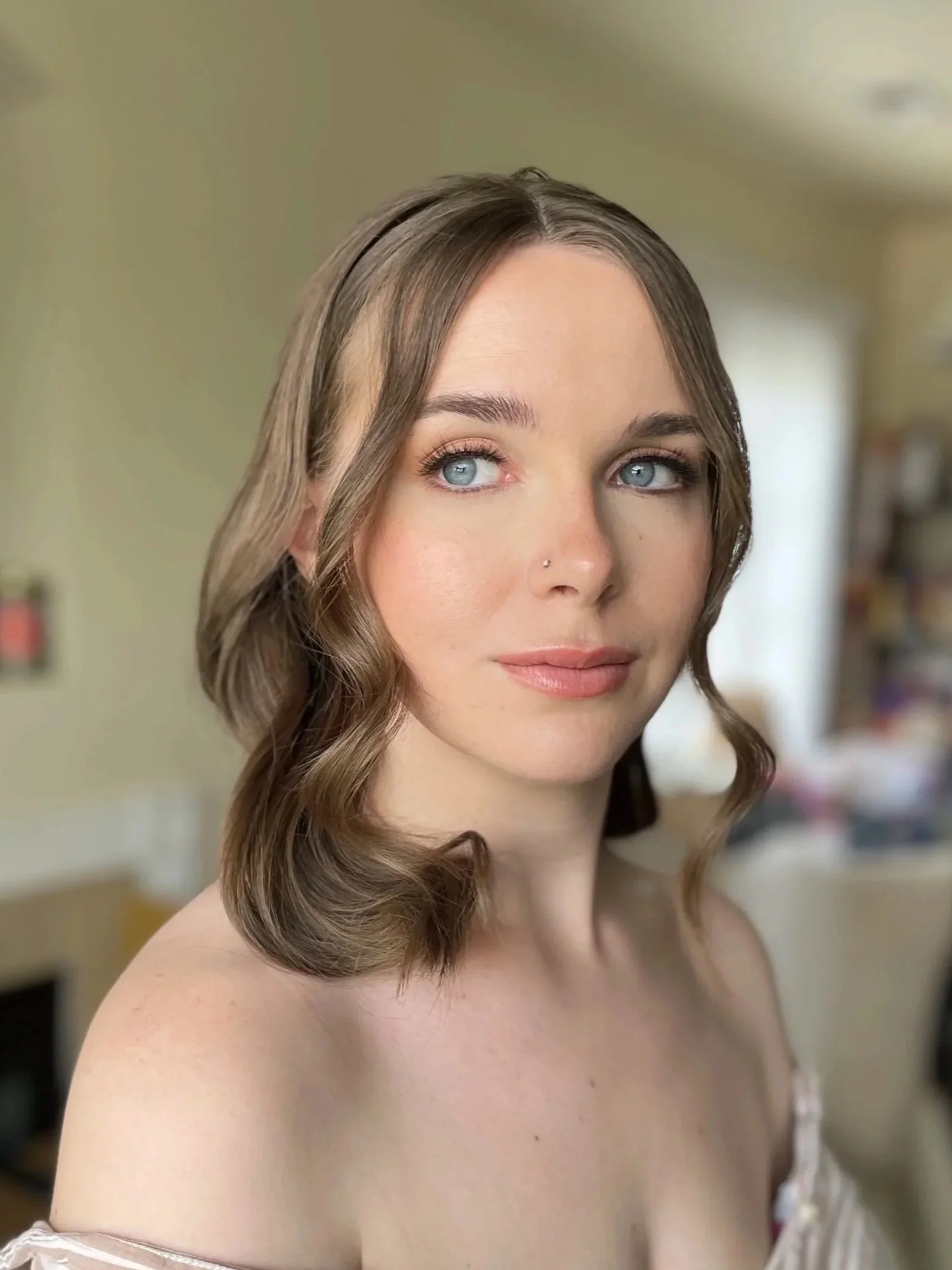 A woman with light brown, wavy hair and blue eyes looking to the side, with a natural makeup look, in a softly lit indoor setting.