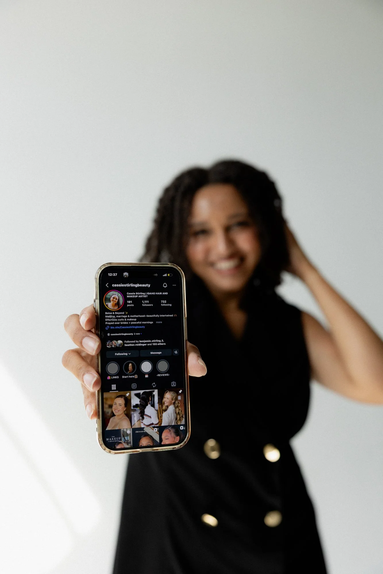 A woman in a black dress holding a smartphone displaying a makeup artist's social media profile, with a plain white background.