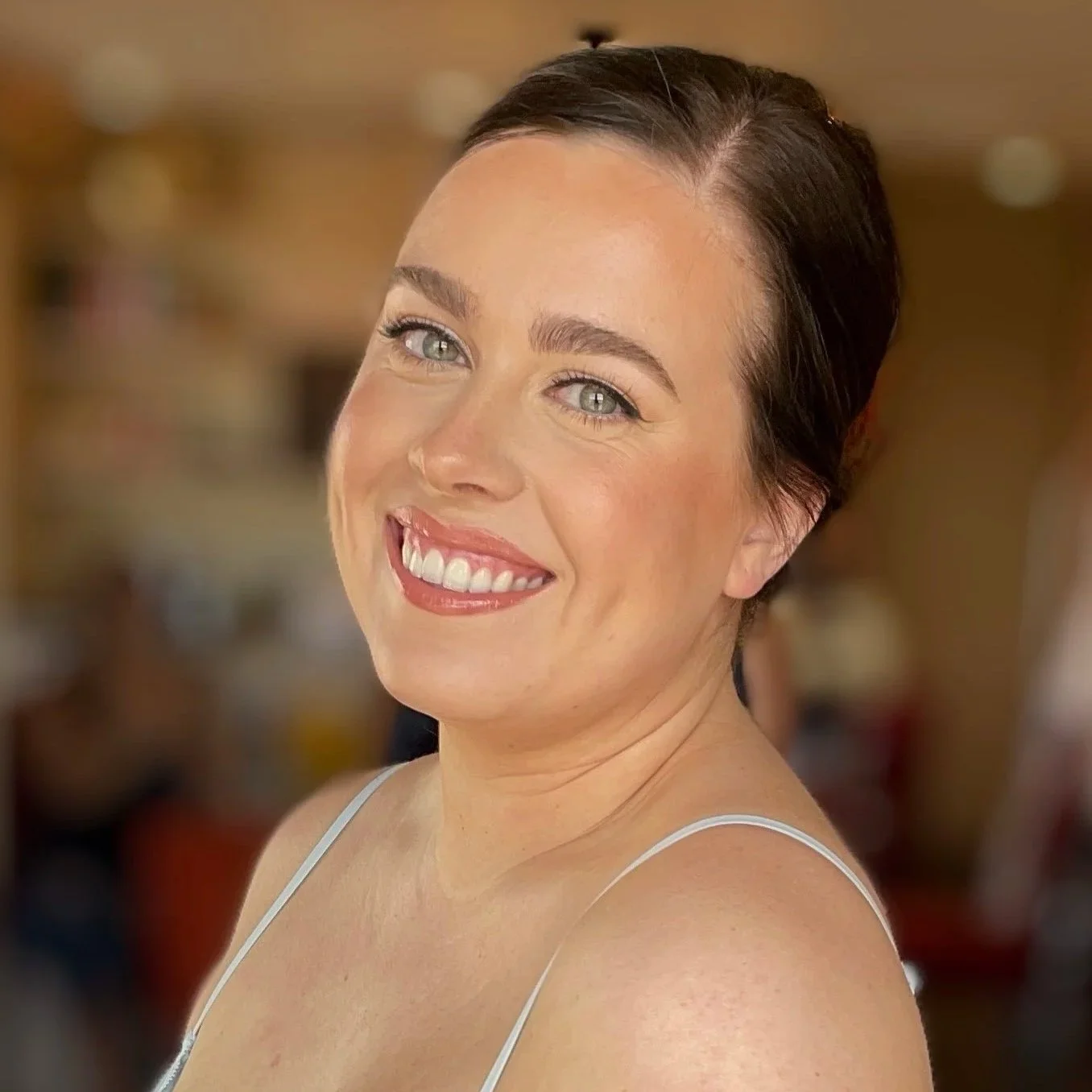 Close-up portrait of a smiling woman with light skin, dark hair, and blue eyes, wearing a white spaghetti strap top, in an indoor setting.