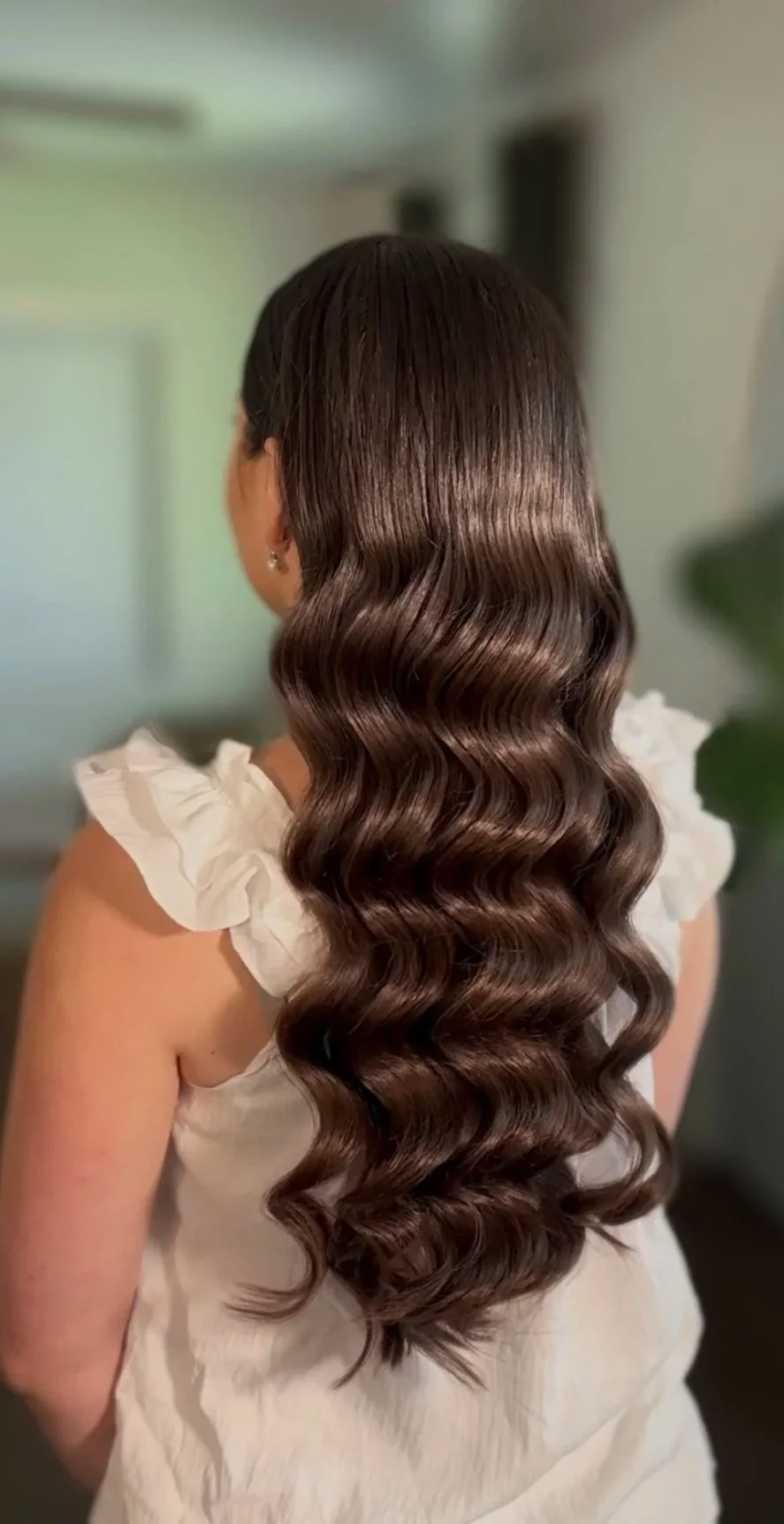 Back of a woman with long, shiny, wavy brown hair wearing a white ruffled top.