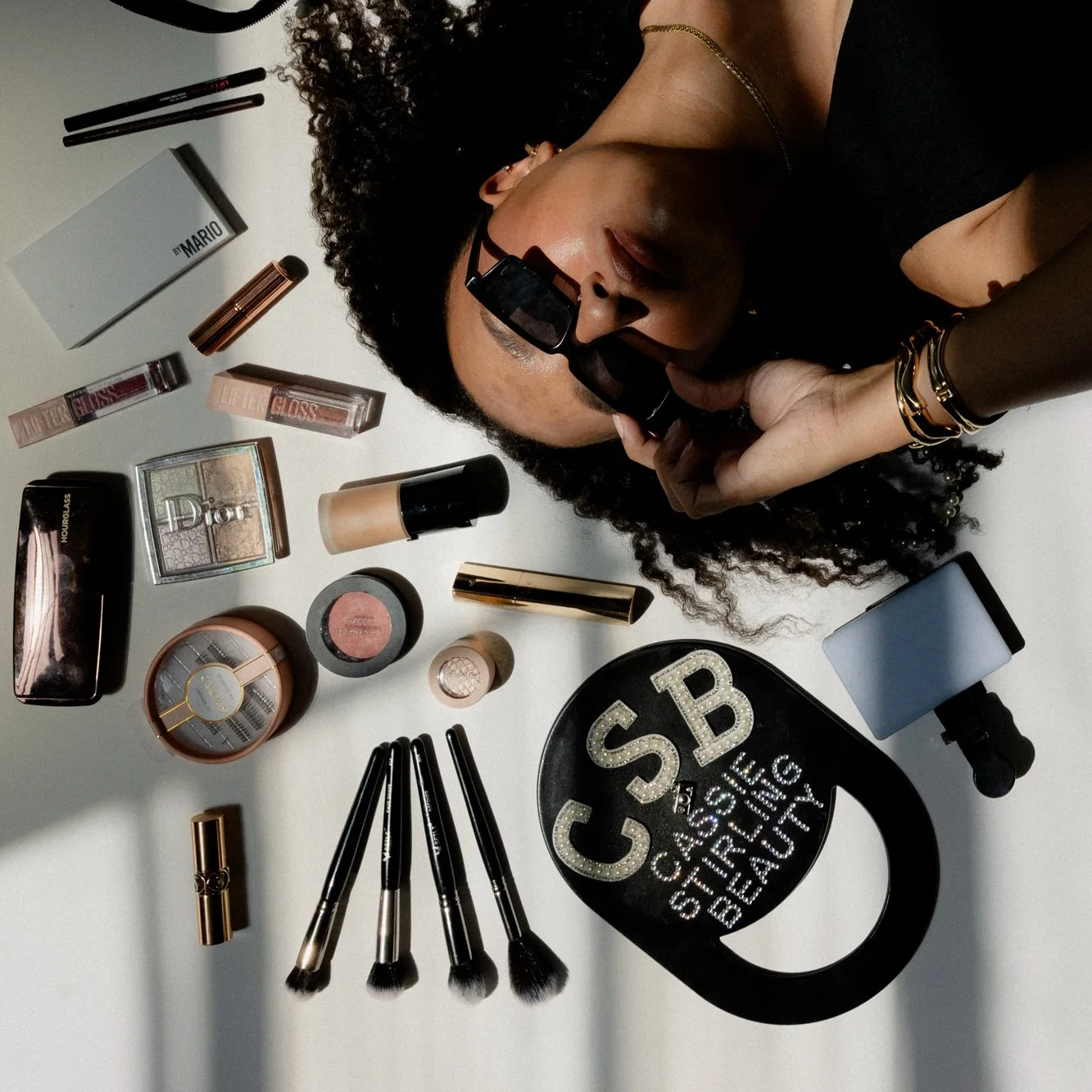 A woman with curly hair, sunglasses, and gold jewelry lying on a white surface surrounded by various makeup products including lipstick, compact powders, brushes, and a black bag with white text.