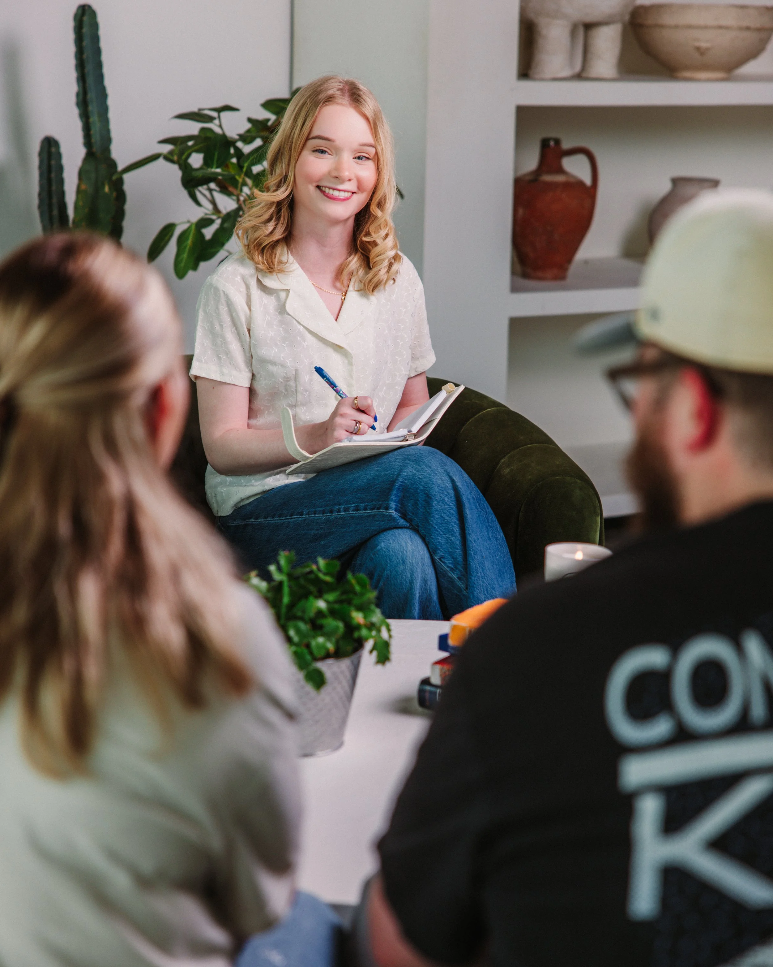 A woman with blonde hair and a white shirt smiling and holding a notepad, sitting on a couch in a cozy room with decorative pottery shelves behind her. Several people are partially visible in the foreground.