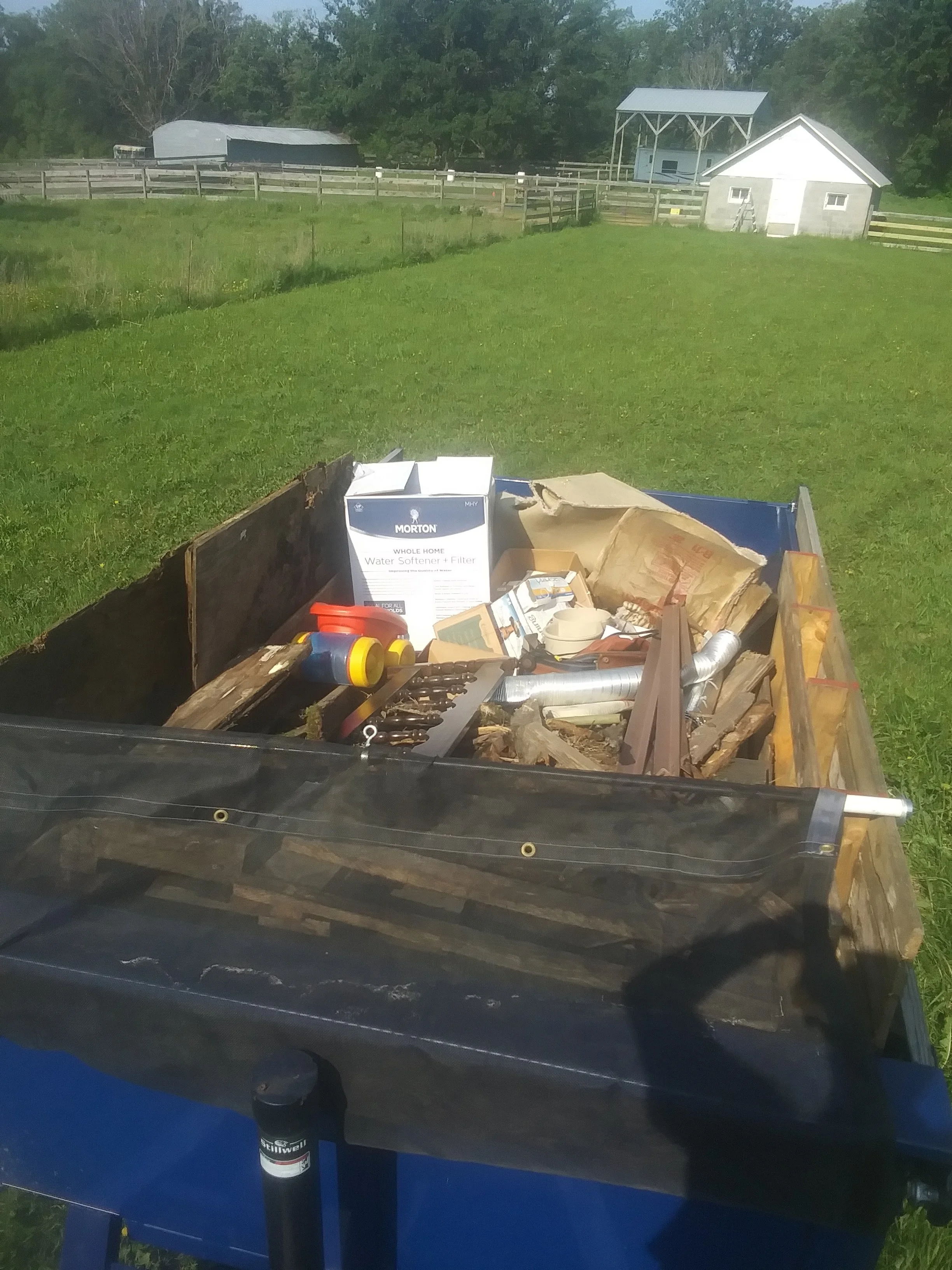 Back of a small utility trailer filled with various tools, a box labeled 'Water Softener + Filter,' and miscellaneous hardware, parked on a grassy field with farm buildings and fences in the background.
