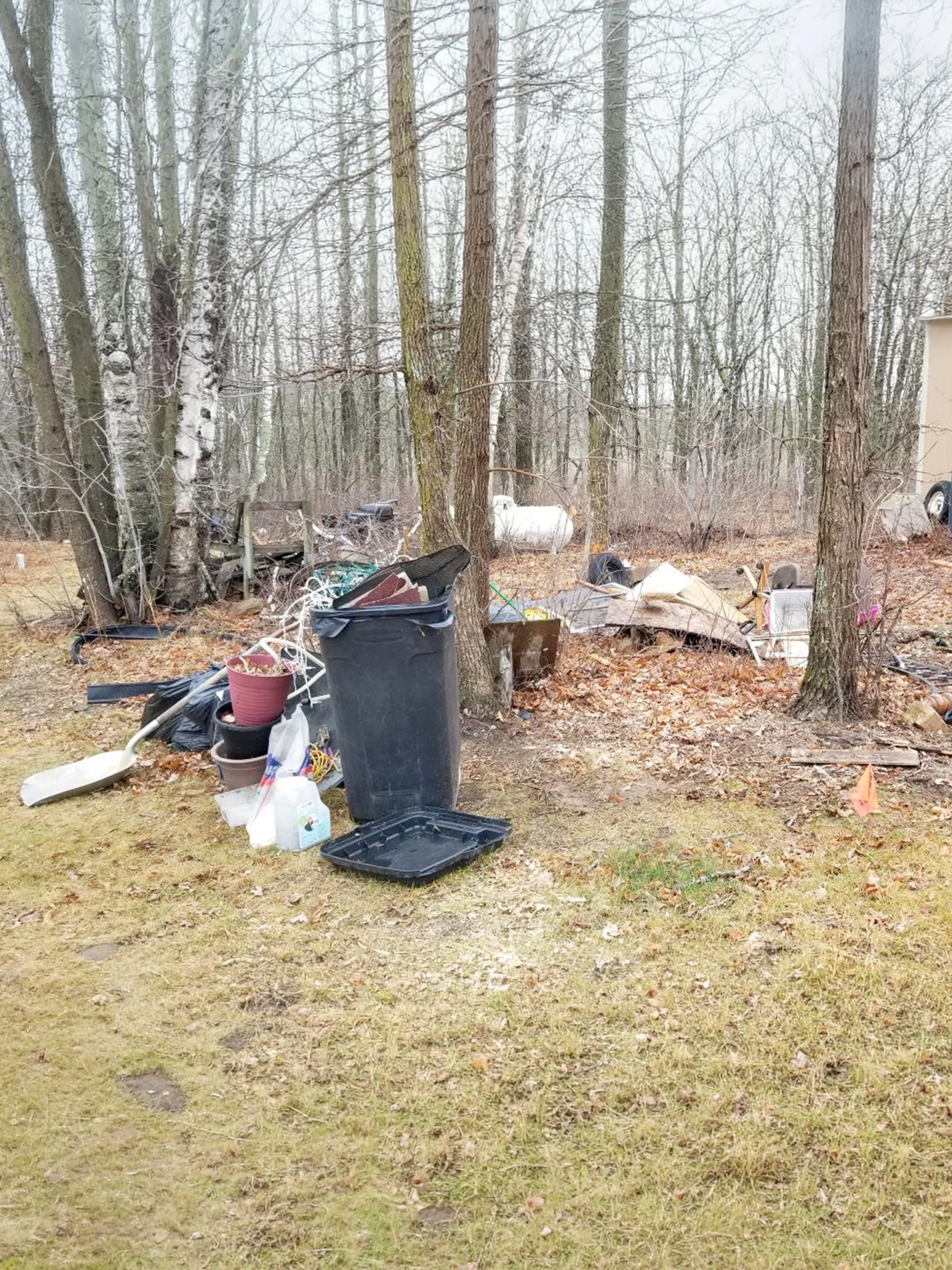 A backyard scene with numerous discarded items including trash cans, tools, and debris scattered among trees and fallen leaves.