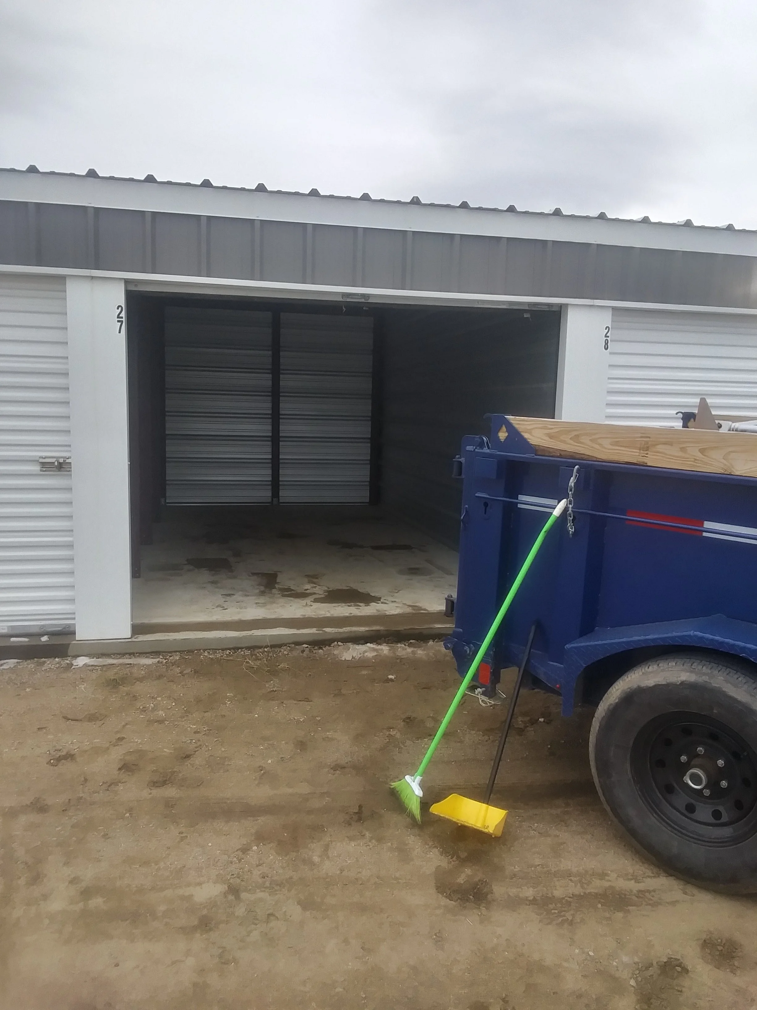 A storage unit with an open garage door, a blue utility cart with wooden top, cleaning broom and dustpan outside on muddy ground, overcast sky above.