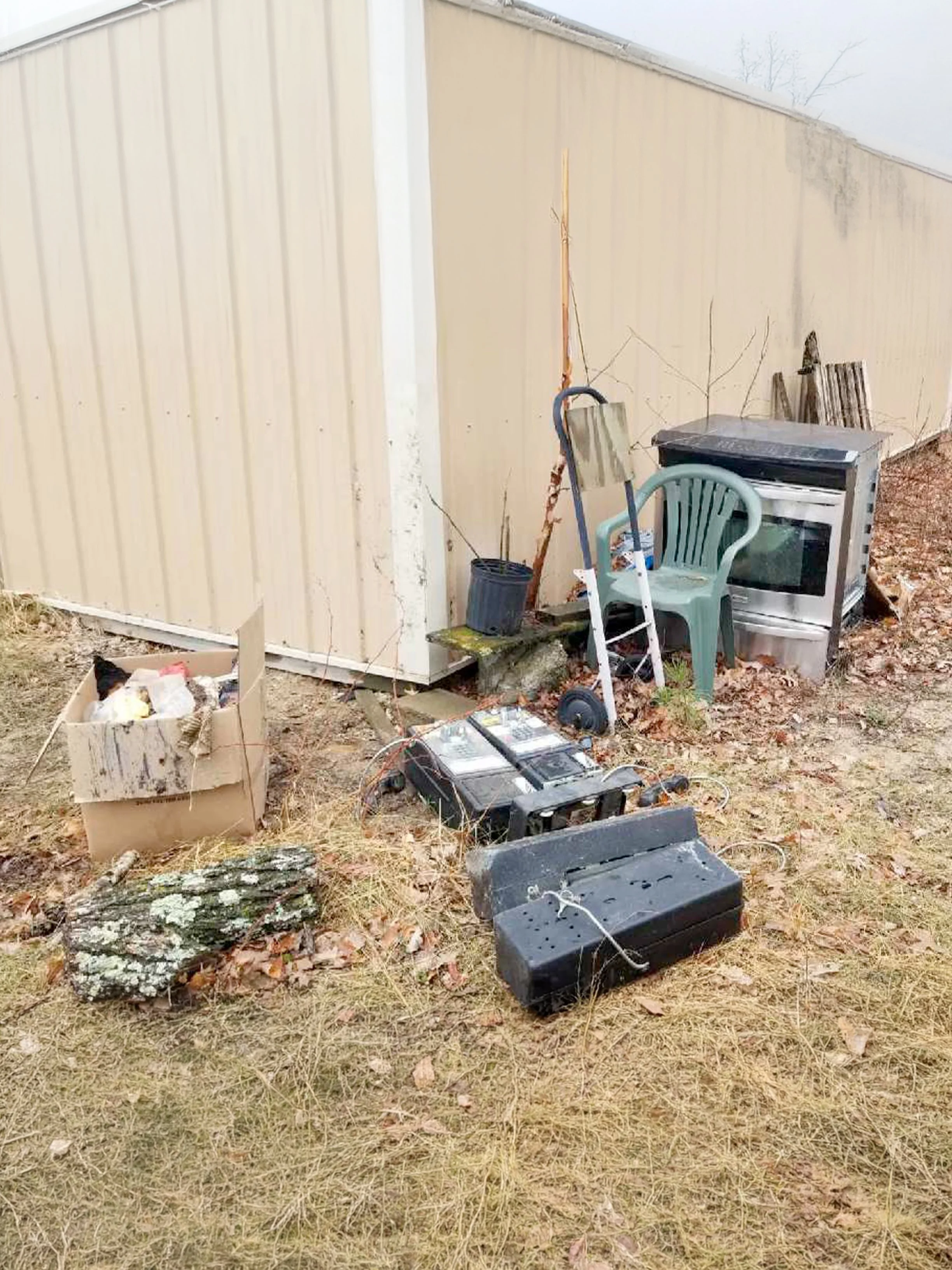 Outdoor scene with discarded electronic equipment, a plastic chair, a small oven, and other miscellaneous items near a beige building wall, with dry grass and fallen leaves on the ground.