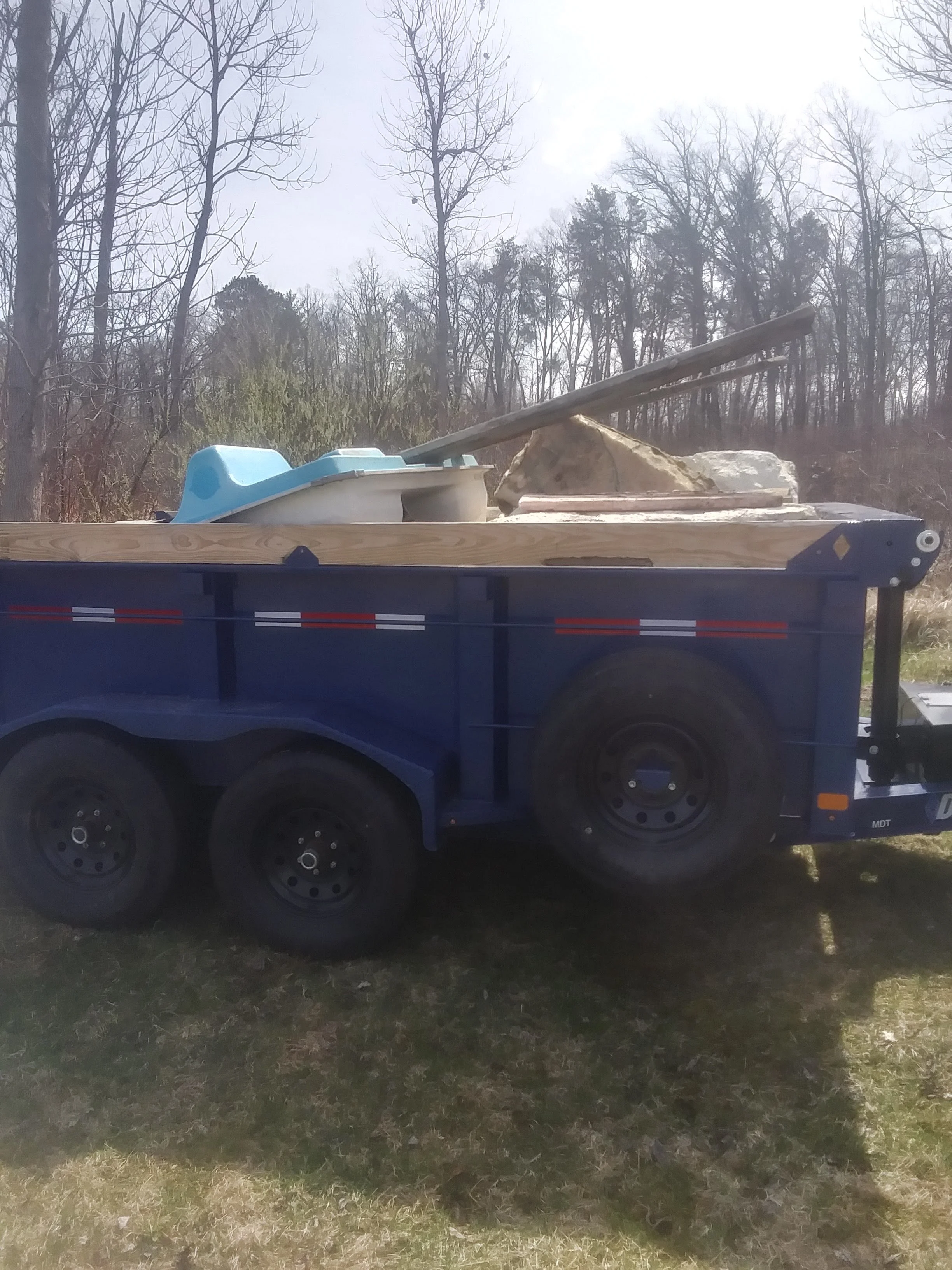 A flatbed trailer carrying construction materials, including a blue baby plastic slide, a large stone, and wooden planks, is parked on grass with leafless trees in the background on a partly cloudy day.