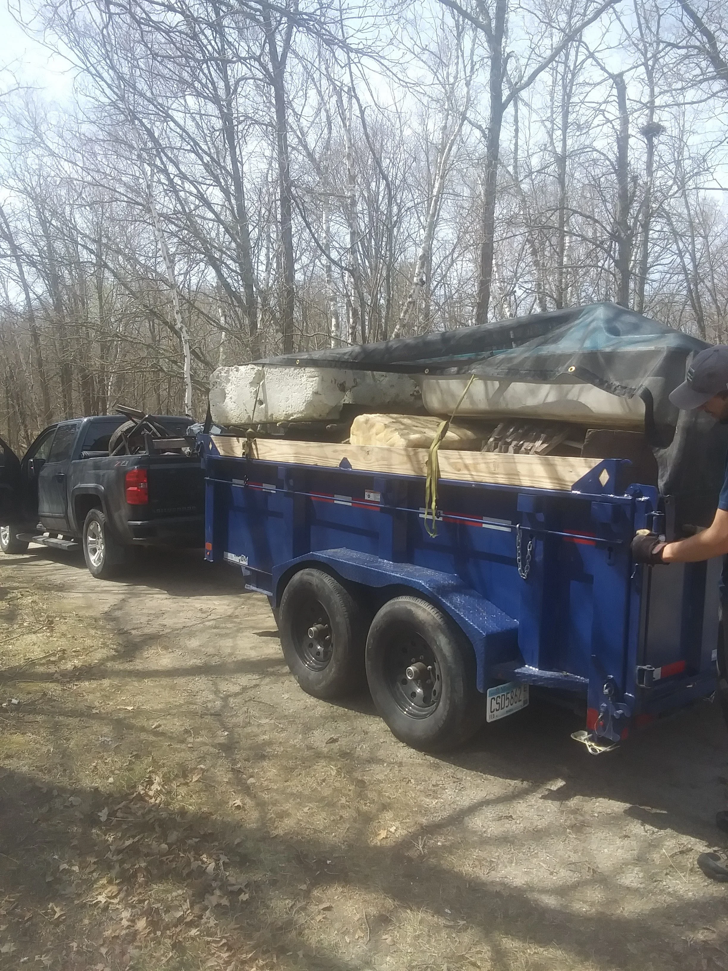 Pickup truck with a blue trailer carrying large foam blocks, parked outdoors on a dirt surface with leaf debris, surrounded by leafless trees.