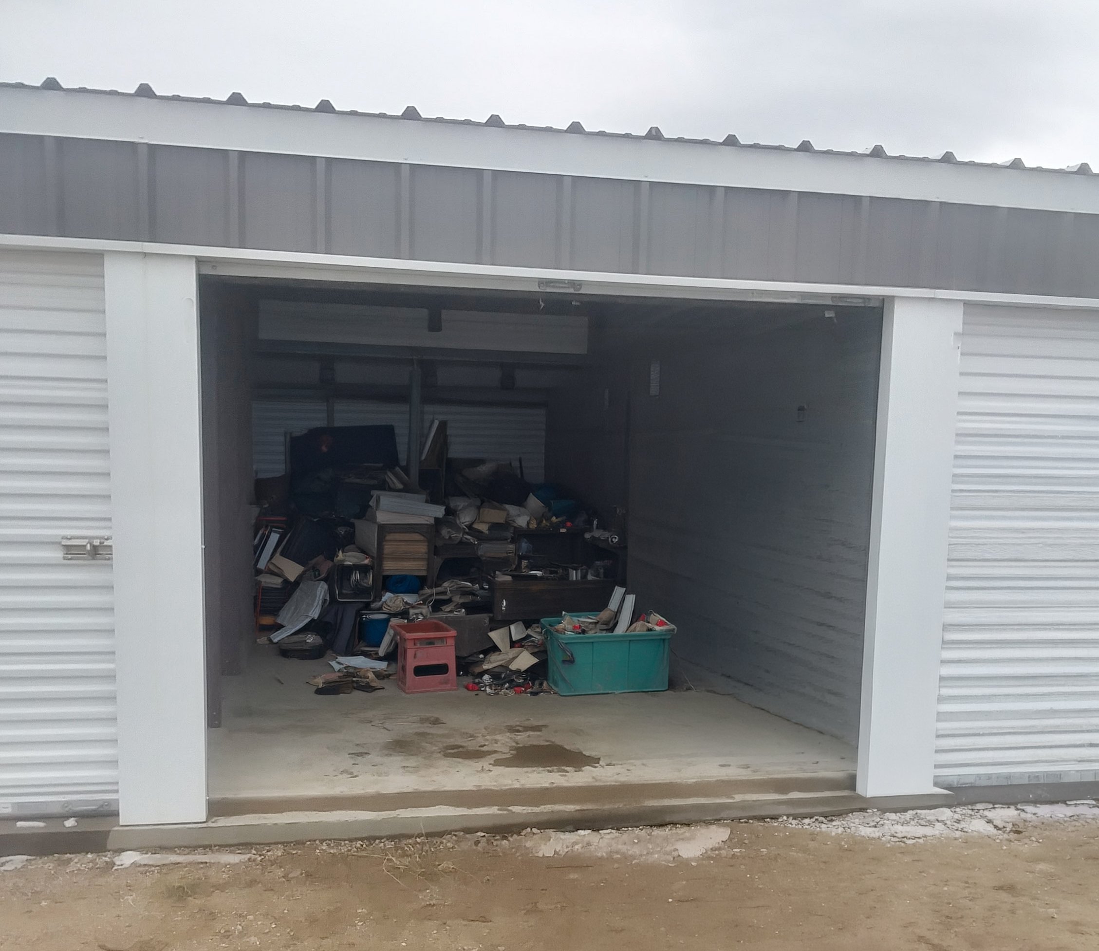 A garage with a partially open door showing clutter inside, including books, boxes, and miscellaneous items. The garage is attached to a building with white siding, and there is a concrete driveway and dirt ground in front.