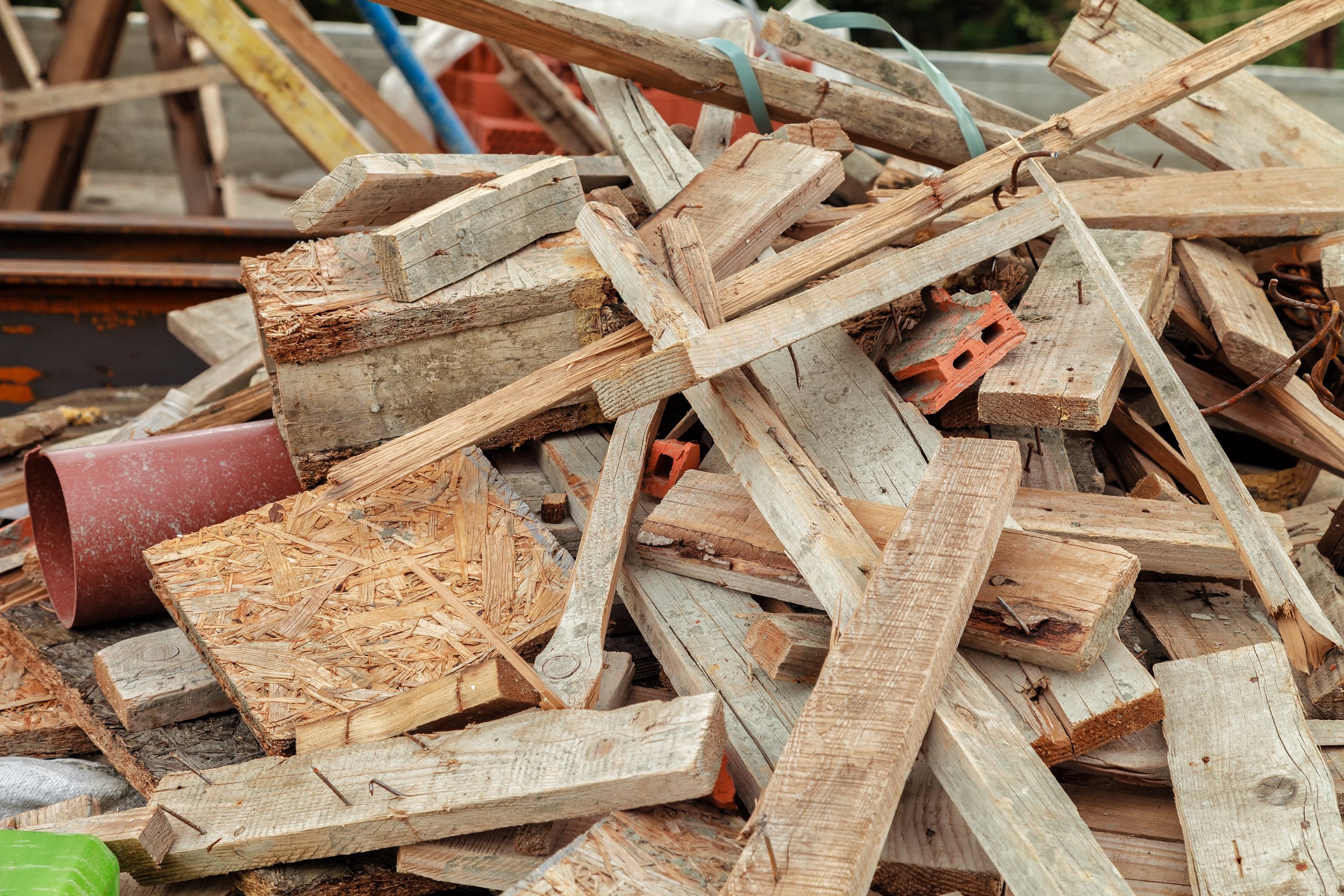 A pile of discarded wooden scrap pieces, some with rusty nails, with construction debris like a red brick and a green plastic item visible in the foreground.