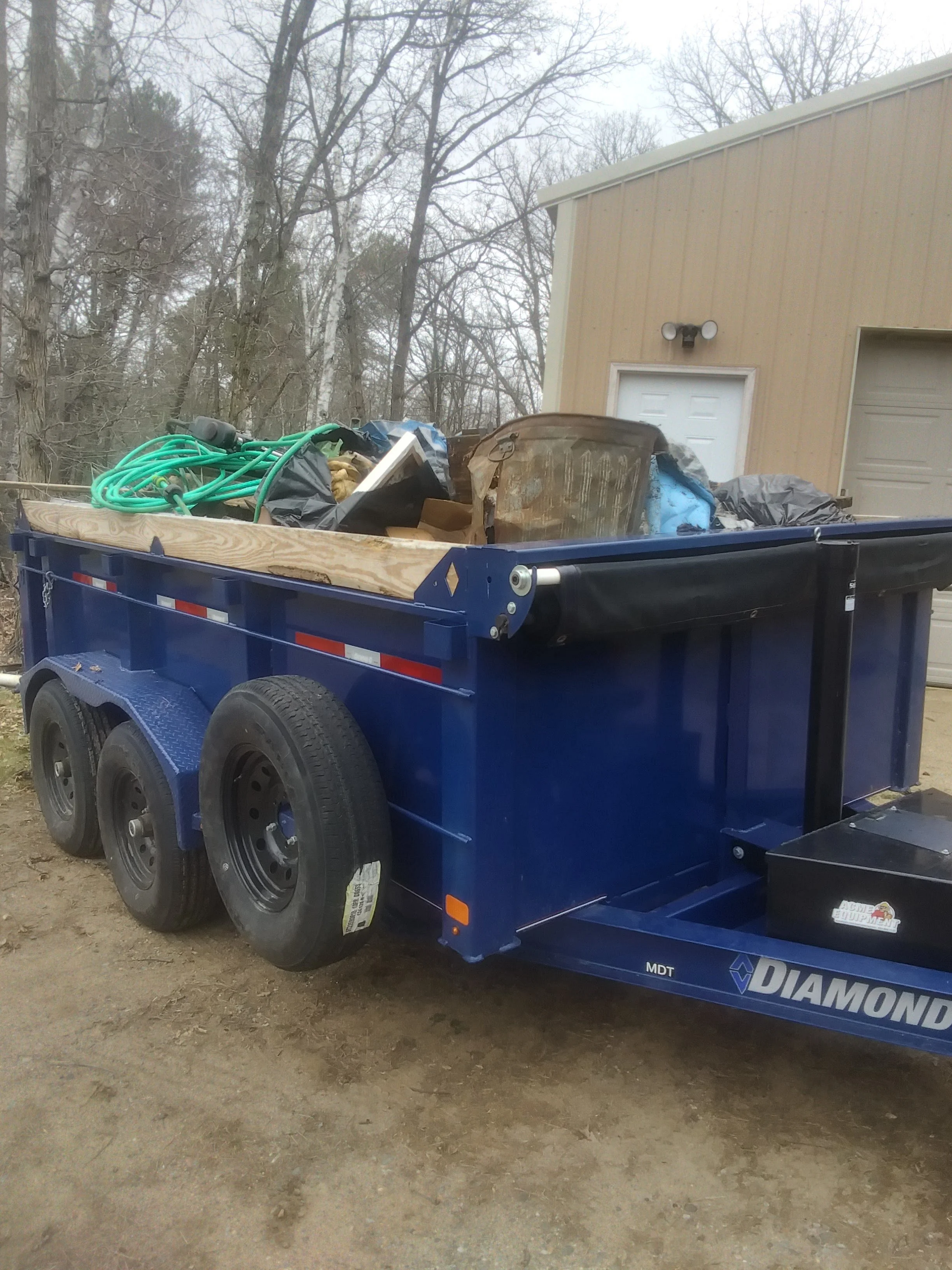 A blue trailer filled with construction debris, including hoses, a chair, and bags, parked on a dirt surface with a beige building and leafless trees in the background.