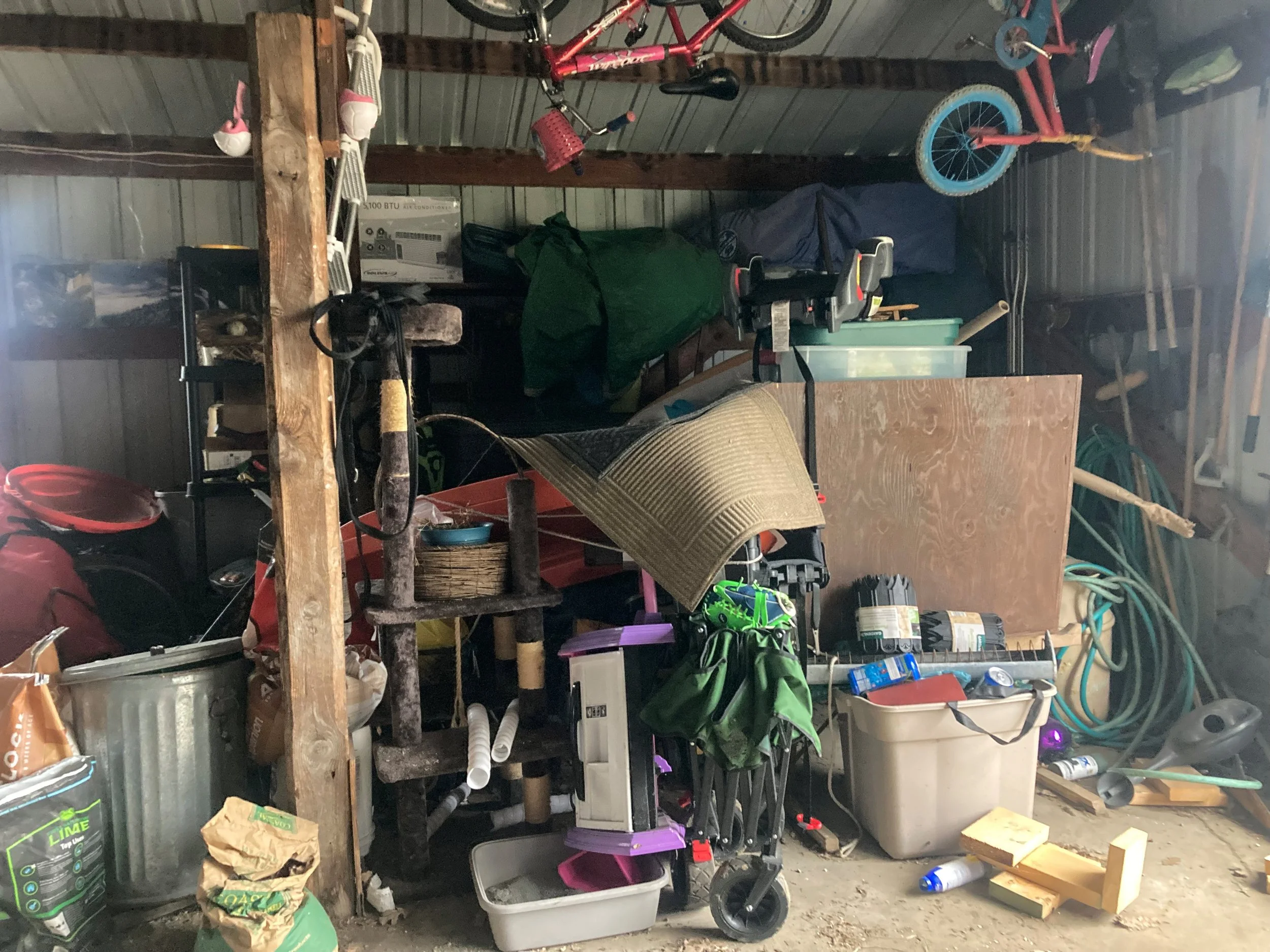 A cluttered garage storage area with household items and bicycles hanging on the wall. There are various containers, tools, and miscellaneous objects stacked and scattered throughout.