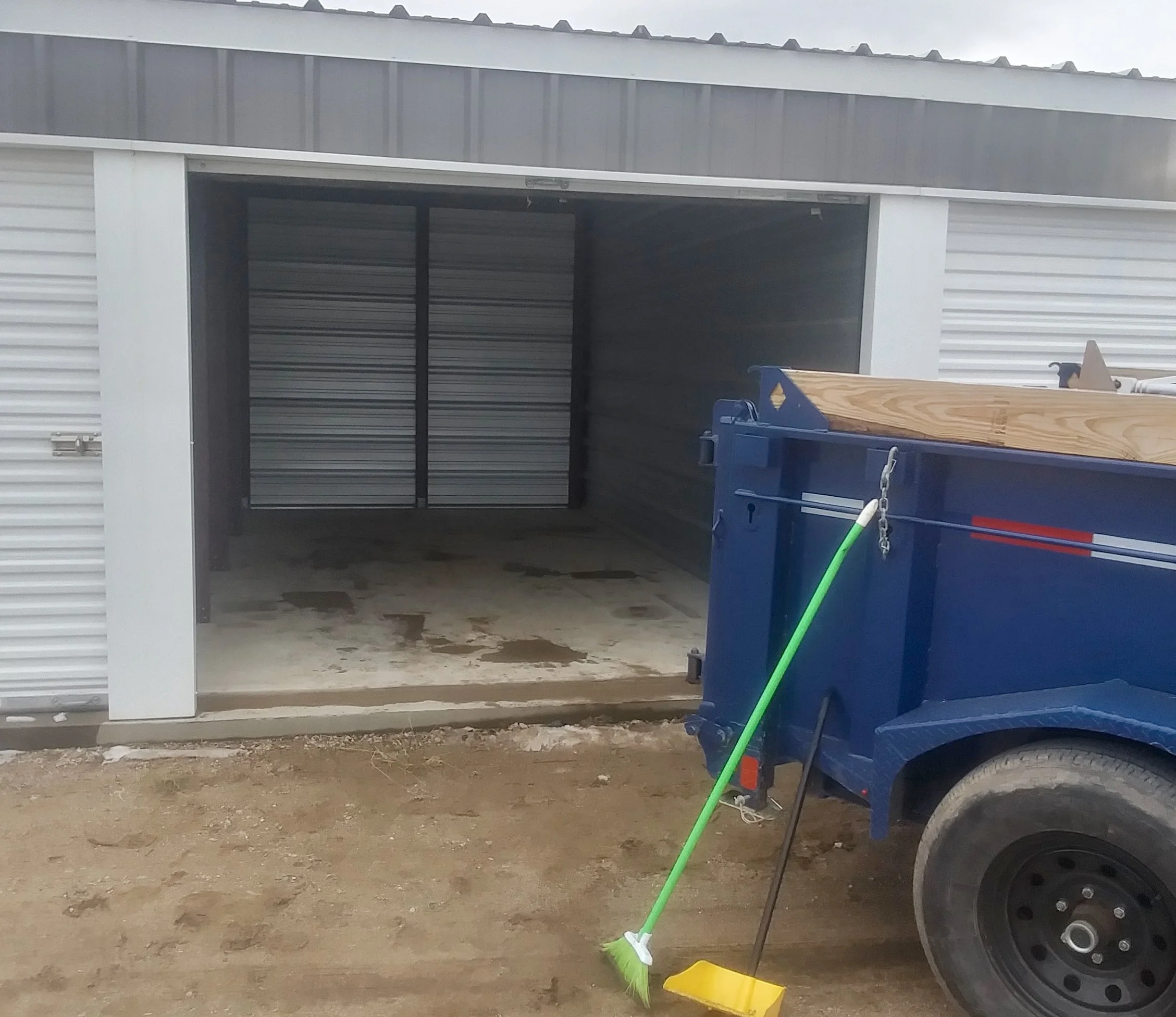 Storage unit with a roll-up door, part of a white metal building, storage bins, a broom, and a green and yellow dustpan on the ground outside.