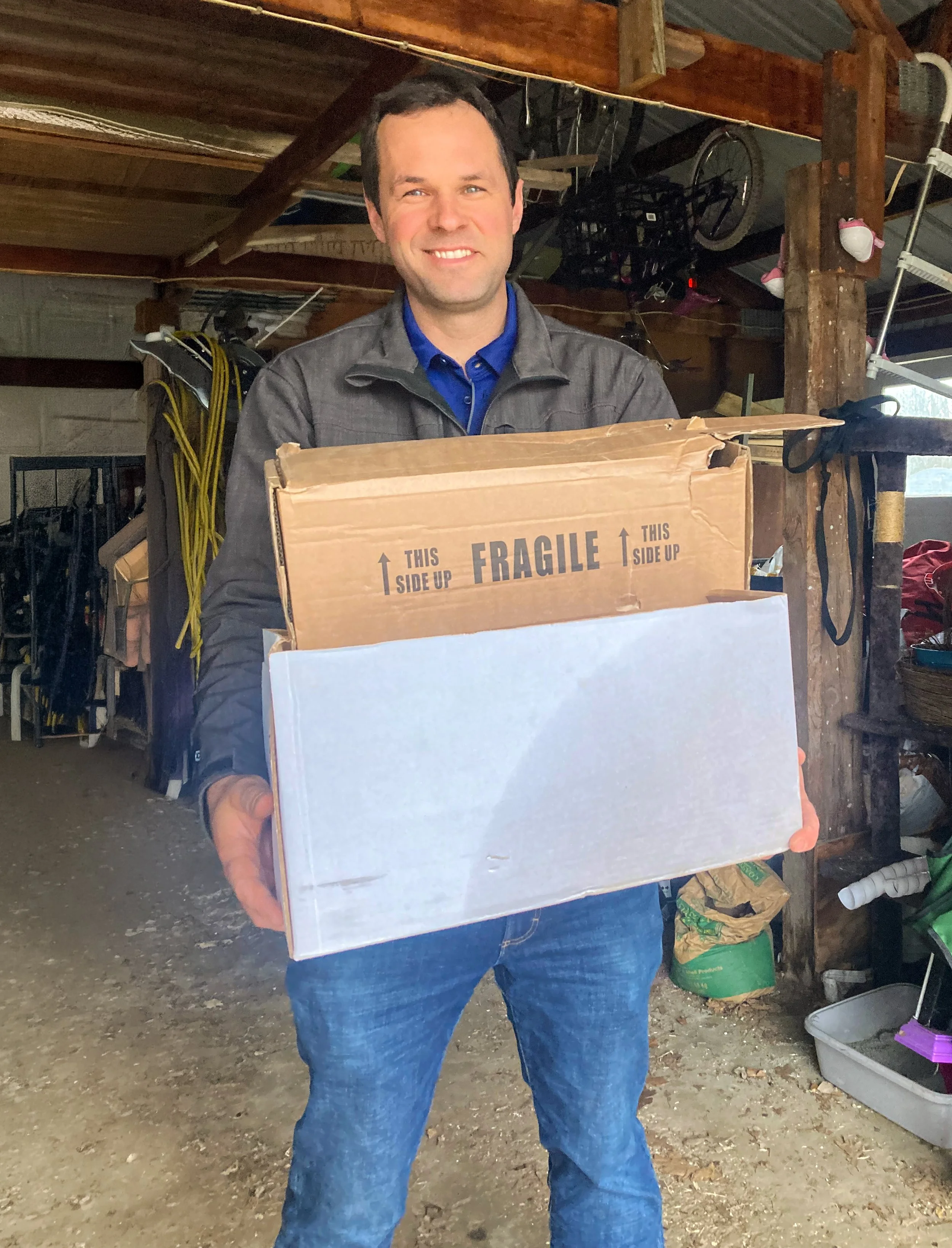 A man wearing a gray jacket and blue shirt holding a large cardboard box, standing inside a rustic shed or barn with wooden beams and various tools and equipment in the background.