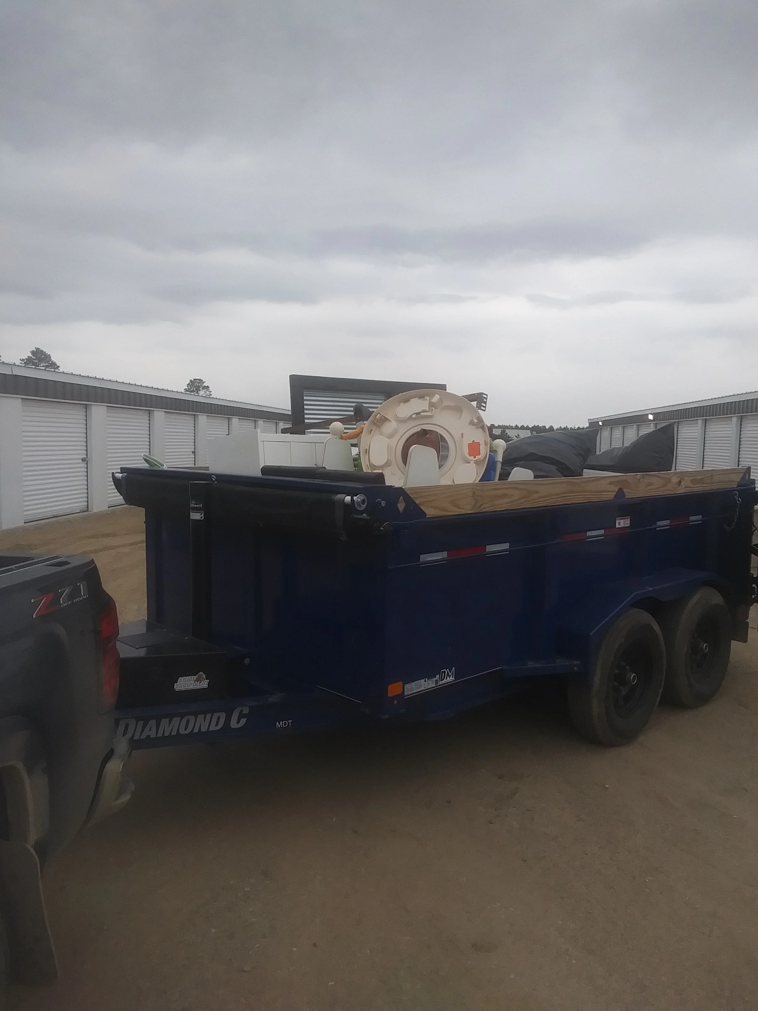 A blue utility trailer carrying an old washing machine and other items parked outdoors near storage units under a cloudy sky.