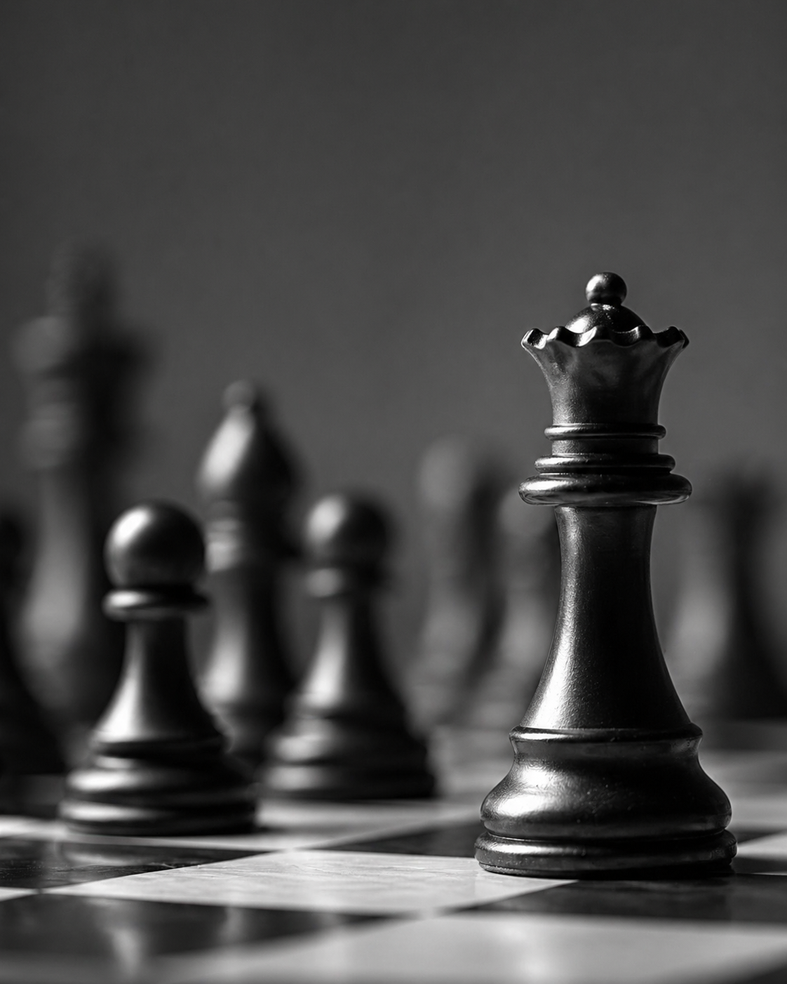 Close-up of a black king chess piece on a chessboard, with several other chess pieces blurred in the background, in black and white.