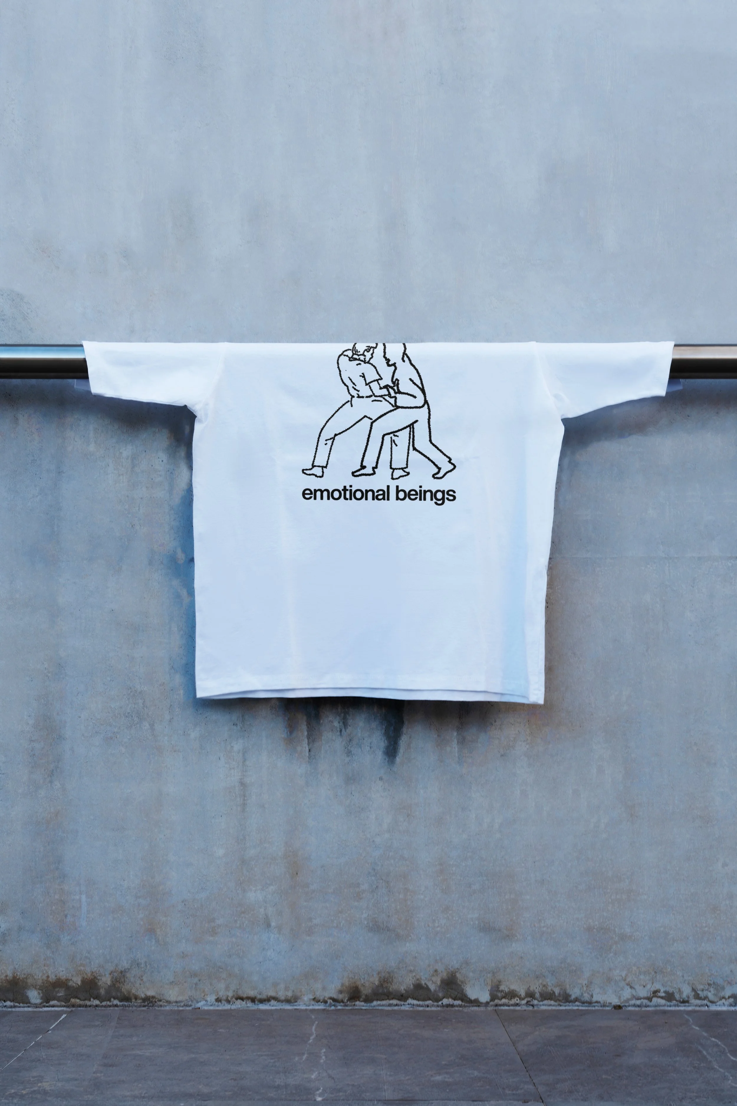A white cloth with a black line drawing of two people sitting and leaning into each other, with the words 'emotional beings' printed below them, hanging on a horizontal metal rod against a textured gray wall.