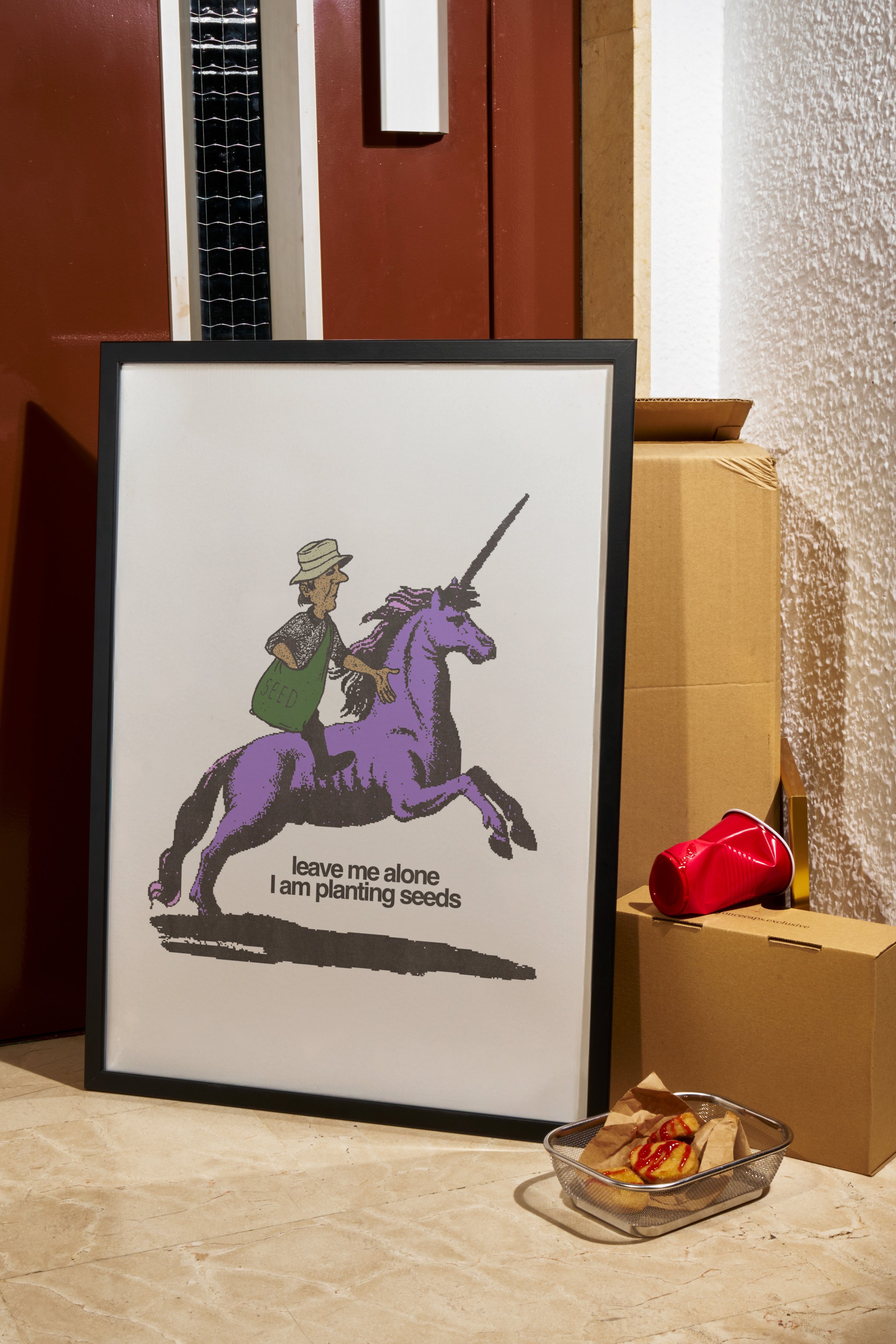 A framed illustration of a person riding a purple unicorn, with the text "leave me alone I am planting seeds" underneath, placed on the floor in a room with a beige marble floor. Behind it, there's a cardboard box, a red object, and a small wire bask