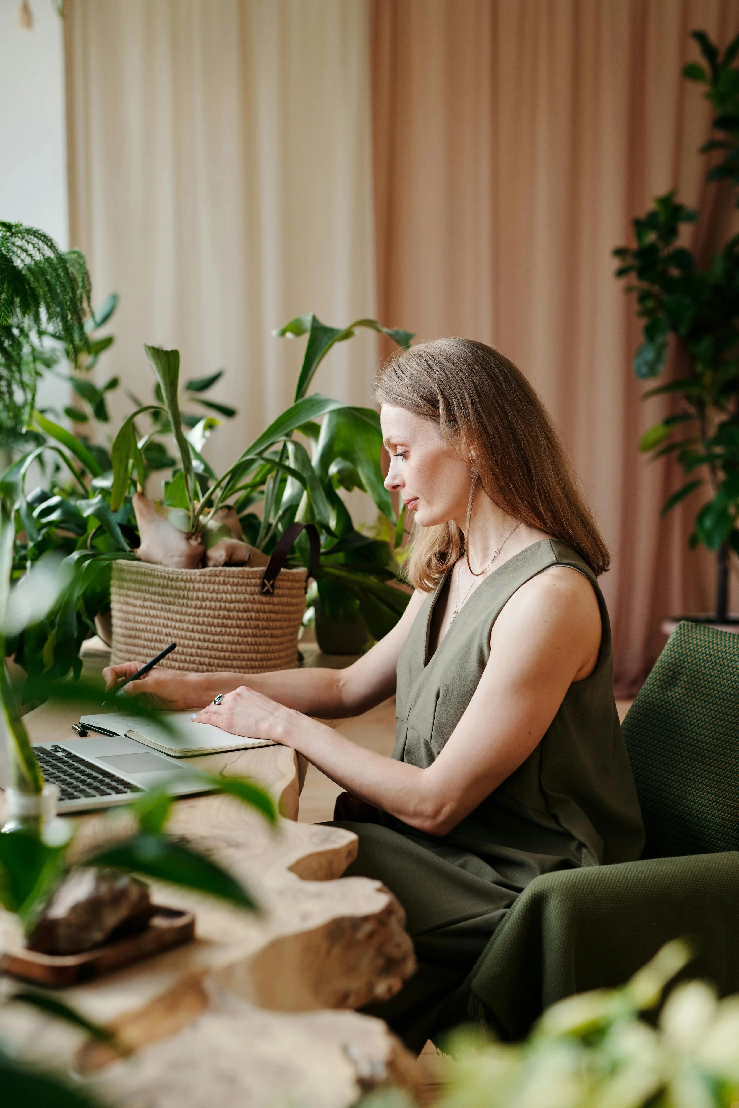 First Last who is a therapist in Asheville, NC writing in a notebook at a wooden desk with a laptop, surrounded by indoor plants.
