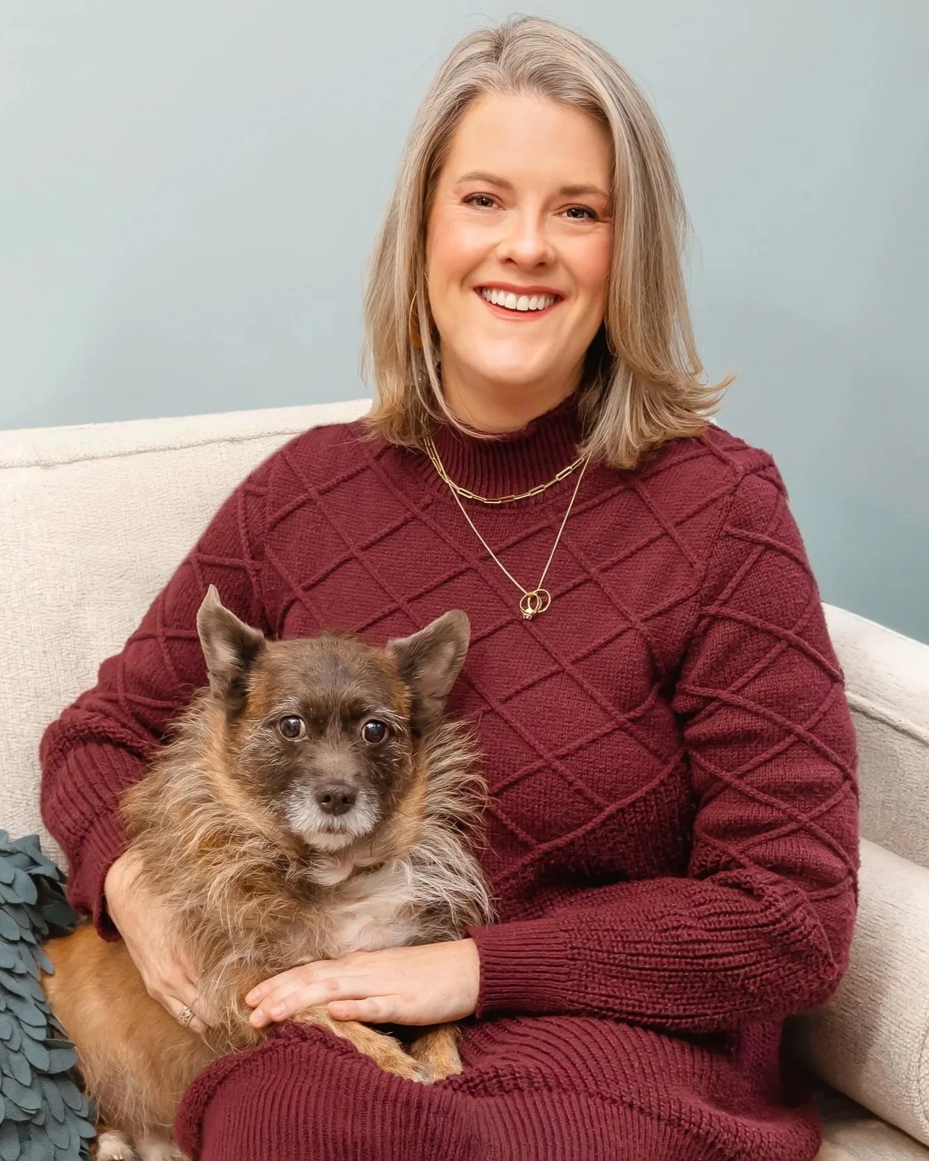 A woman with shoulder-length blonde hair, wearing a maroon sweater and gold jewelry, sitting on a beige couch holding a small brown and tan dog with fluffy fur. The woman is smiling at the camera against a light blue wall.