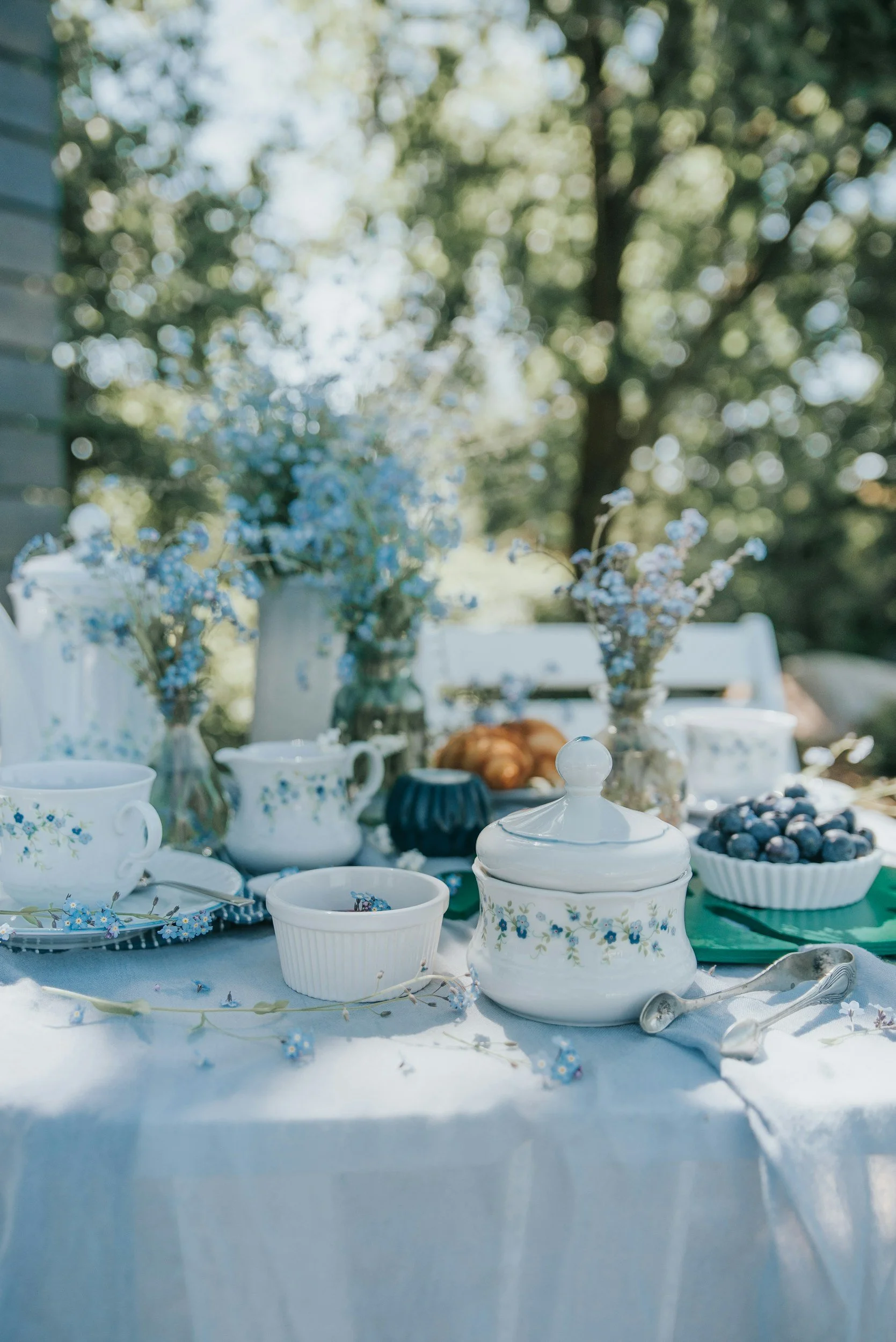 A table set for outdoor tea with vintage china teacups and bowls, a sugar bowl with a floral pattern, blueberries in a bowl, and small flowers scattered on a white tablecloth. In the background, there are trees with green leaves and a wooden fence.