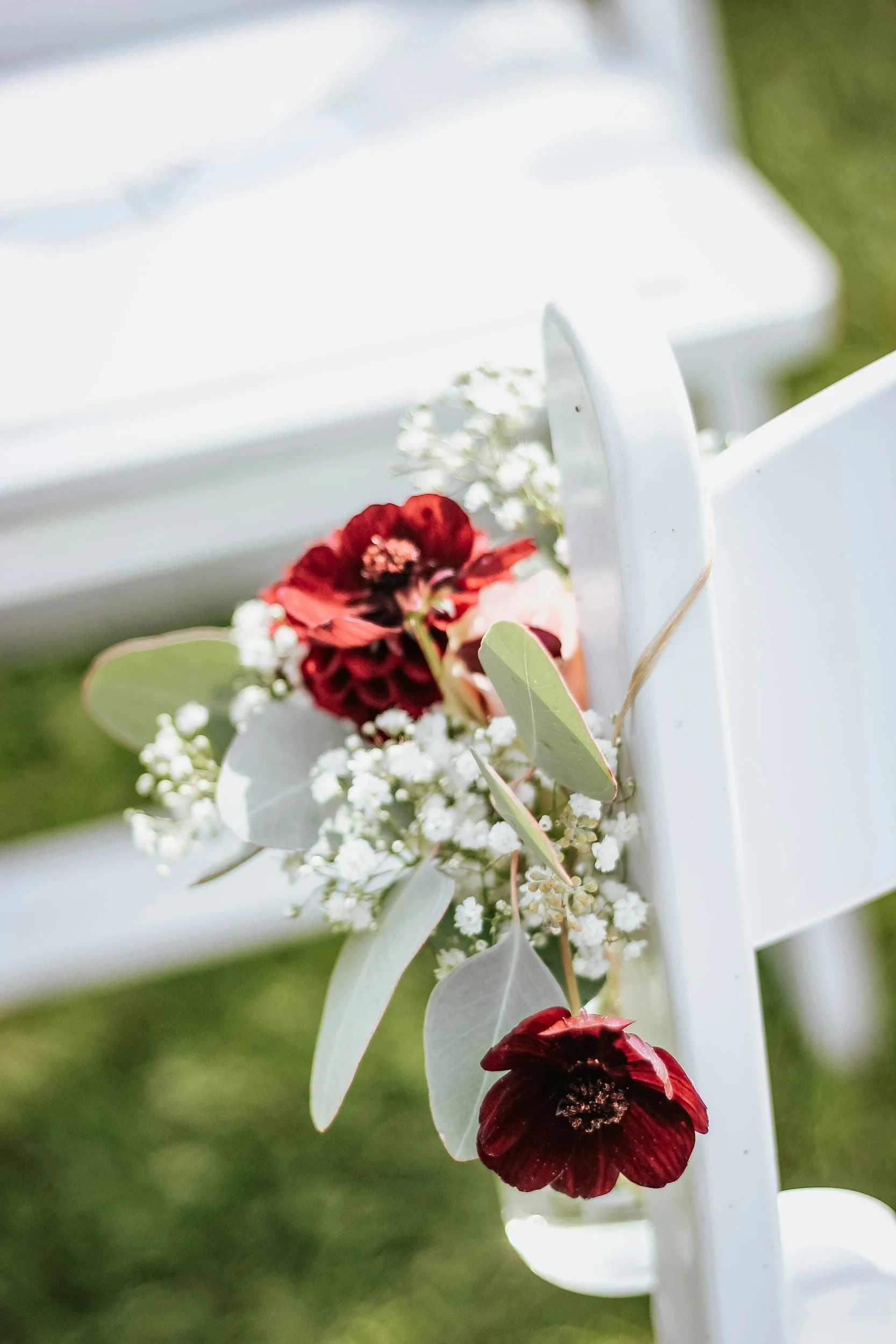 Close-up of a white outdoor chair decorated with red and white flowers, green leaves, and eucalyptus, set on a grassy lawn.