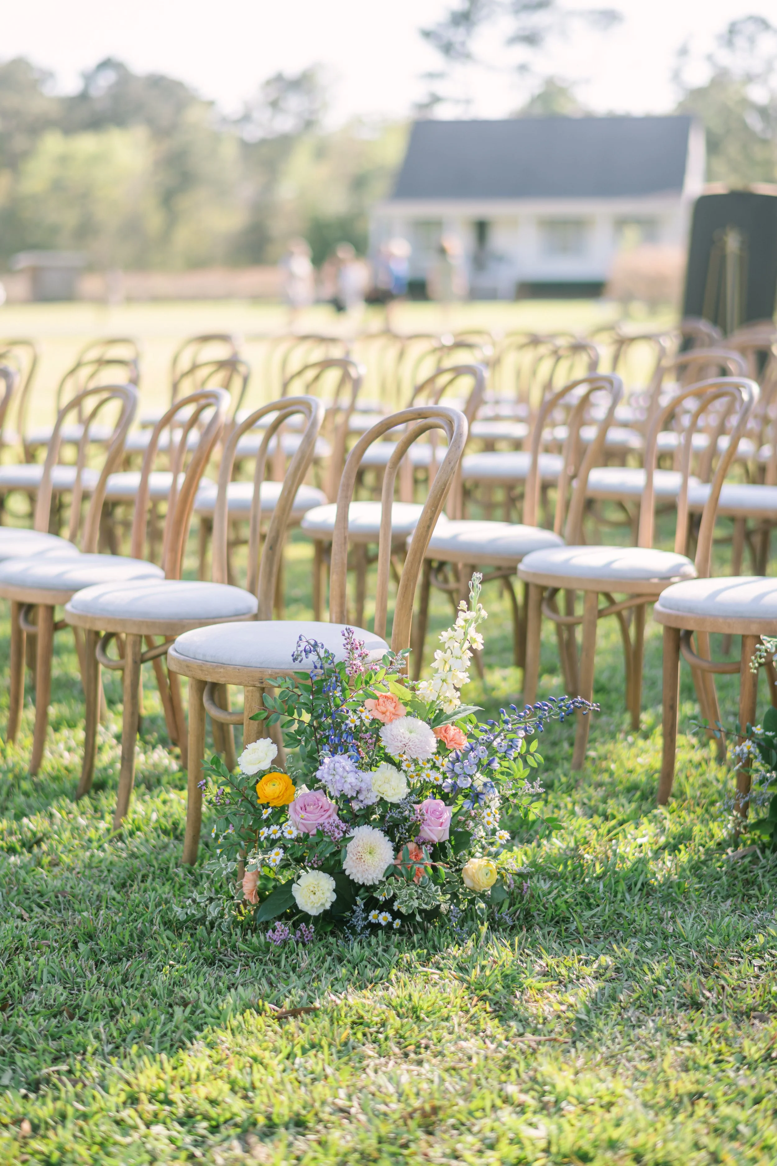 Rows of wooden chairs set up outdoors on green grass with a large flower arrangement in front, with a house and trees in the background, suggesting a wedding or outdoor event.