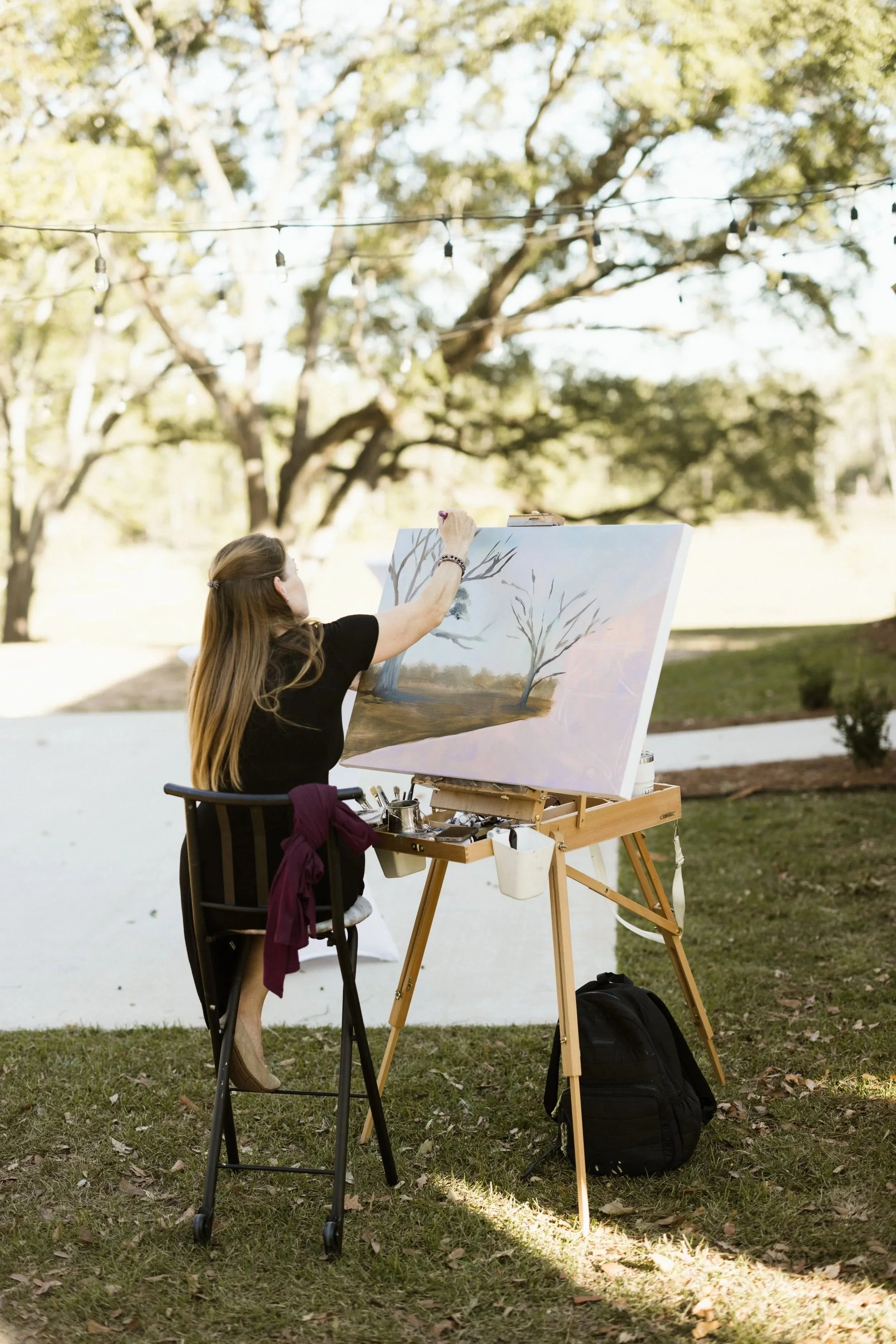 A woman painting outdoors on a large canvas, seated on a chair with art supplies on a wooden easel. She has long hair, is wearing a black top, and is focused on her artwork. There are trees and string lights in the background, suggesting a park or garden setting.