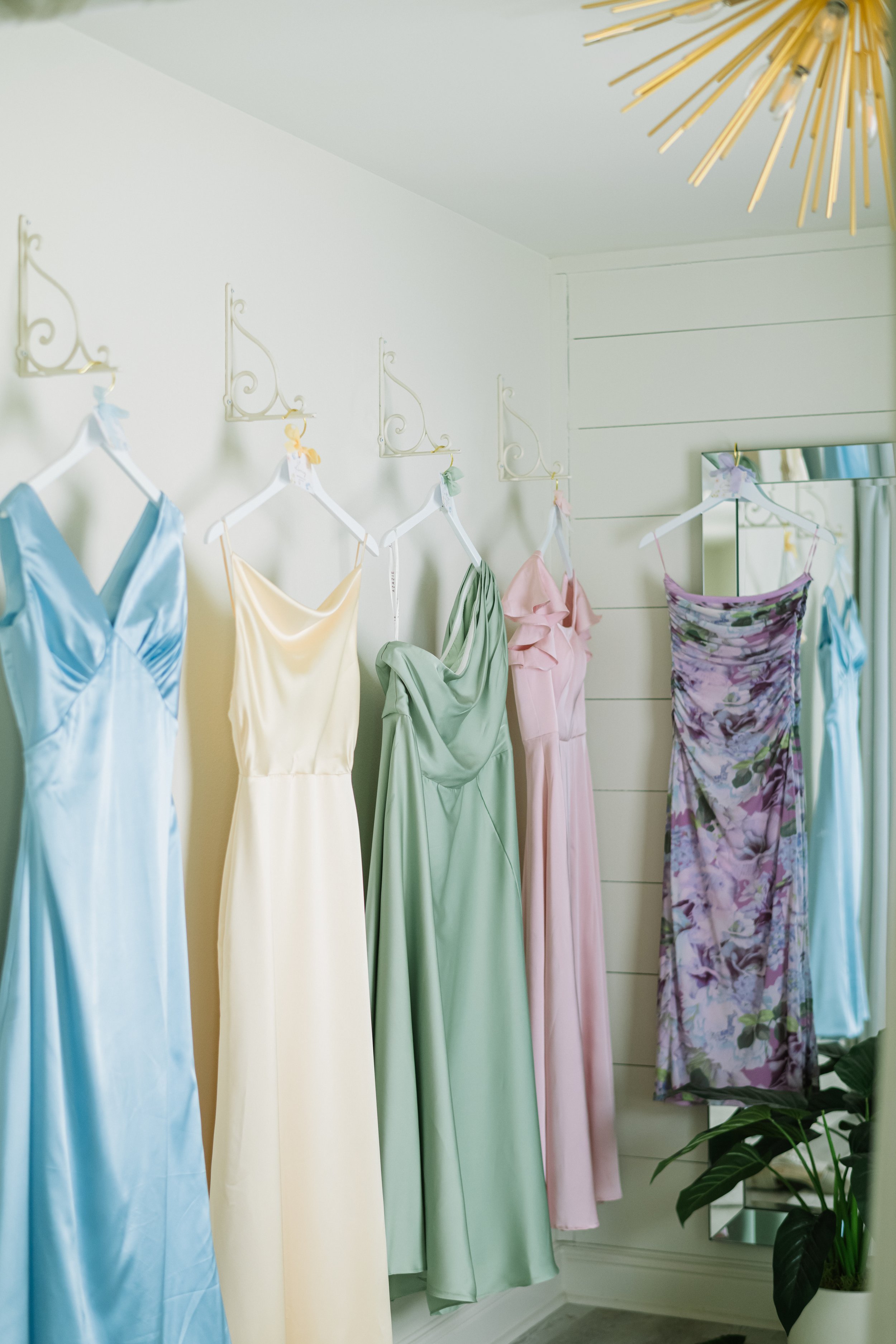 A display of pastel-colored and floral dresses hanging on white hangers and decorative wall shelves in a boutique or dressing room. There are five dresses in shades of blue, yellow, green, pink, and purple, with a mirror and potted plant also visible.
