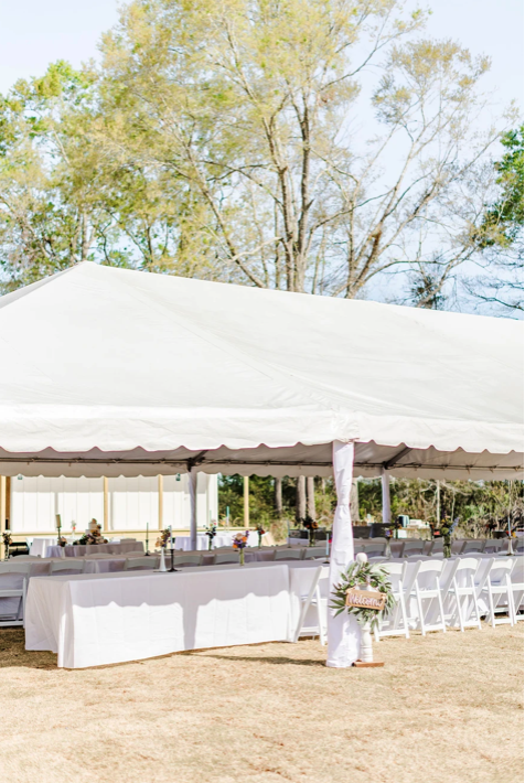 Outdoor event setup with a large white canopy tent, long tables with white tablecloths, and chairs, in a grassy area with trees in the background.