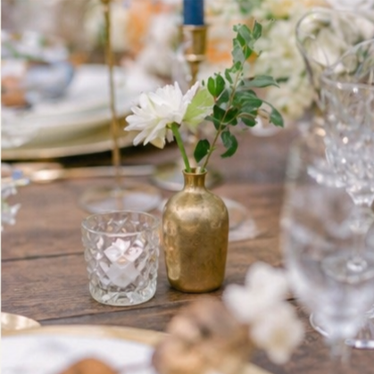 A gold vase with white flowers and greenery on a wooden table, with glasses and tableware in the background.