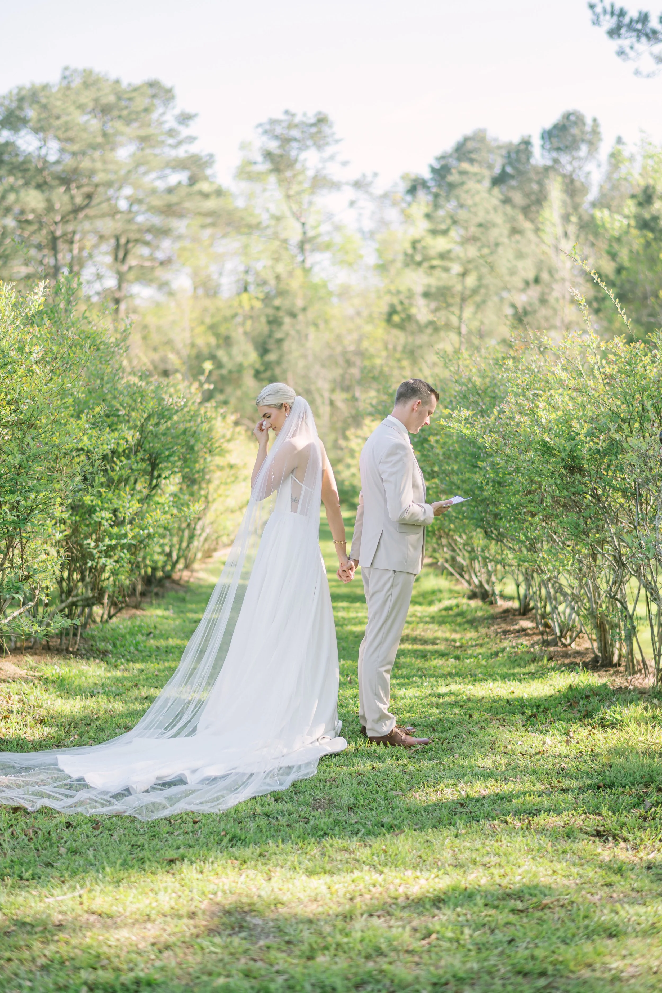 A bride and groom are standing back to back in a lush green outdoor setting, holding hands and looking down, with the bride wearing a white wedding dress and veil and the groom wearing a light-colored suit.